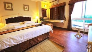 A hotel room with a large bed, a small table, a chair, and a window with curtains. - The Airport Greenery Hotel (Chiang Mai)