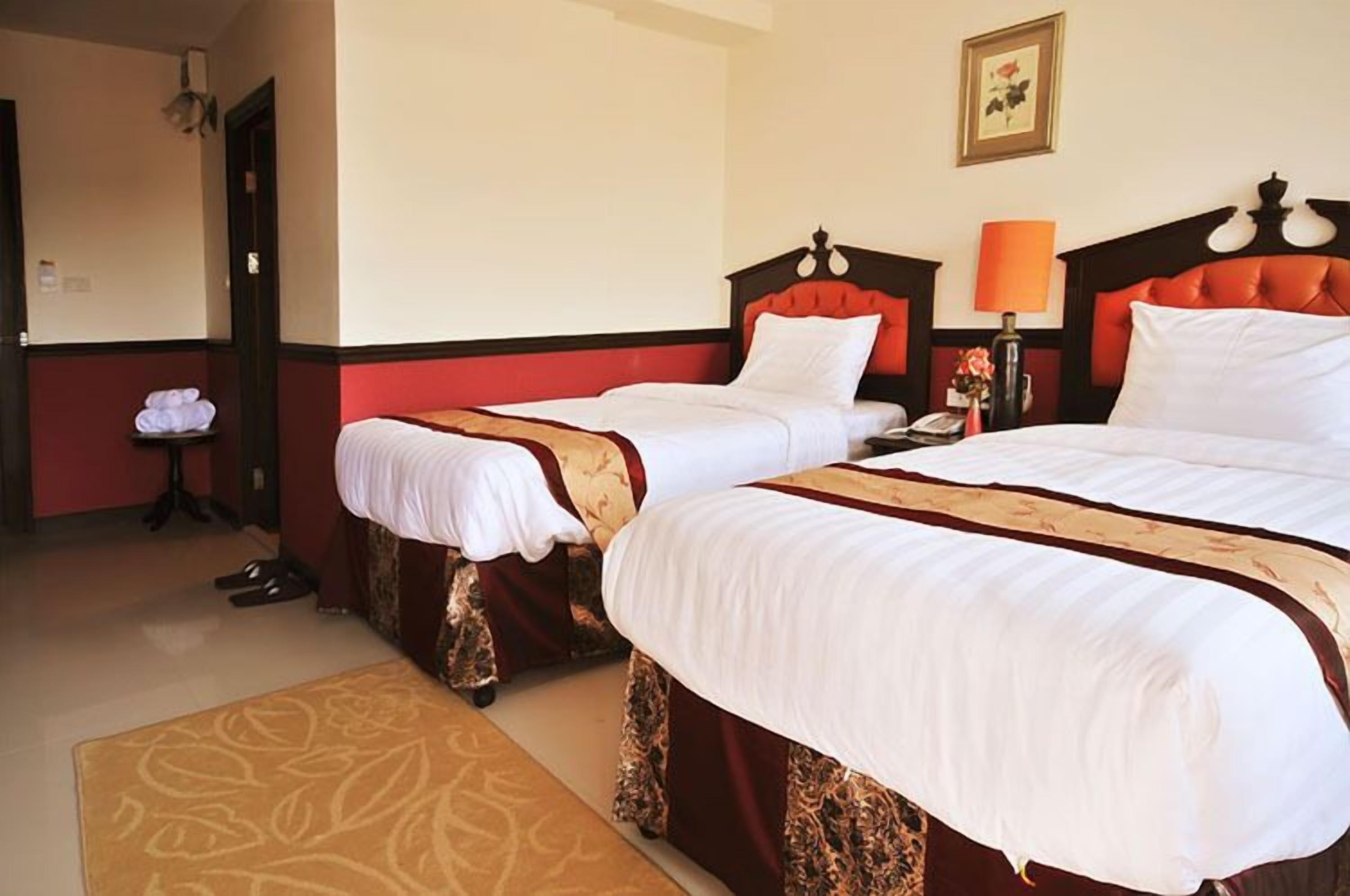 A hotel room with two beds, a bedside table with a lamp, a chair, and a door.