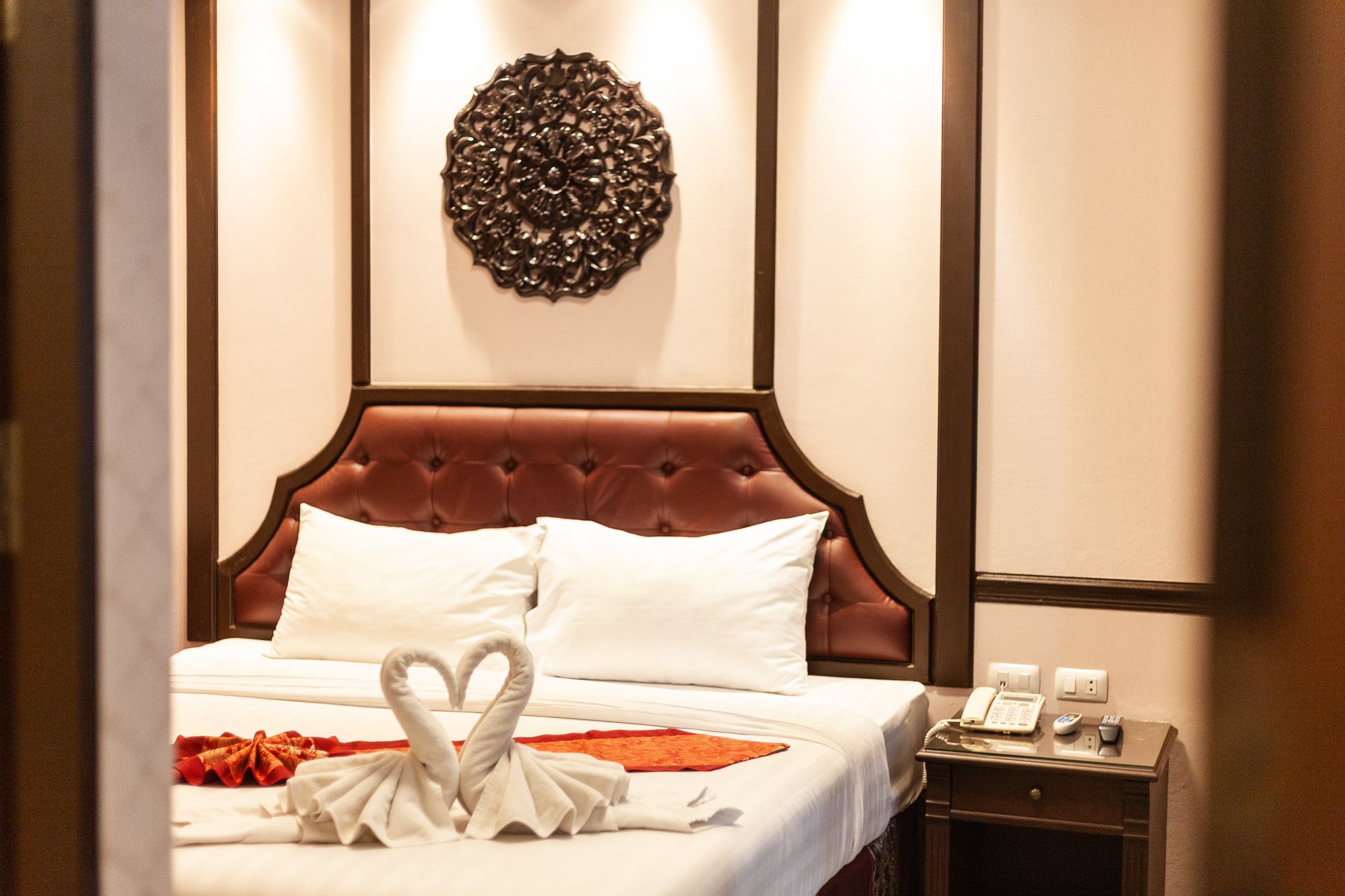 A hotel room with a tufted headboard, white bedding, a decorative wall piece, a bedside table with a phone and a lamp, and a swan-shaped towel arrangement.