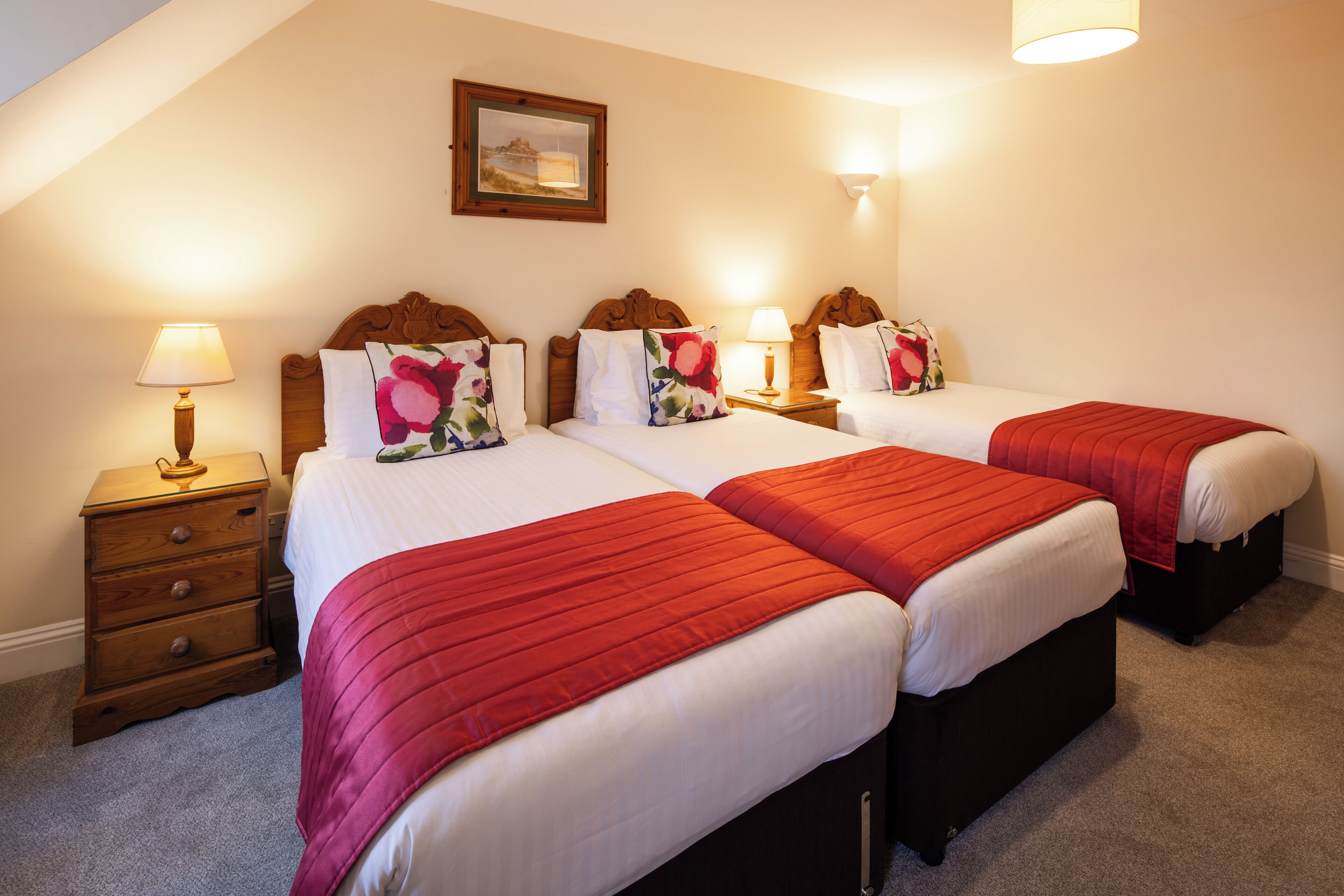 A hotel room with three beds, each with a red bedspread and white linens. There is a wooden nightstand with a lamp on the left side of the room.