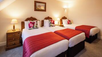 A hotel room with three beds, each with a red bedspread and white linens. There is a wooden nightstand with a lamp on the left side of the room.