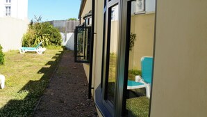 A narrow pathway leading to a building with large windows and a small garden area with a chair and a table. - The Uplands Serviced Apartments (St. Helier)