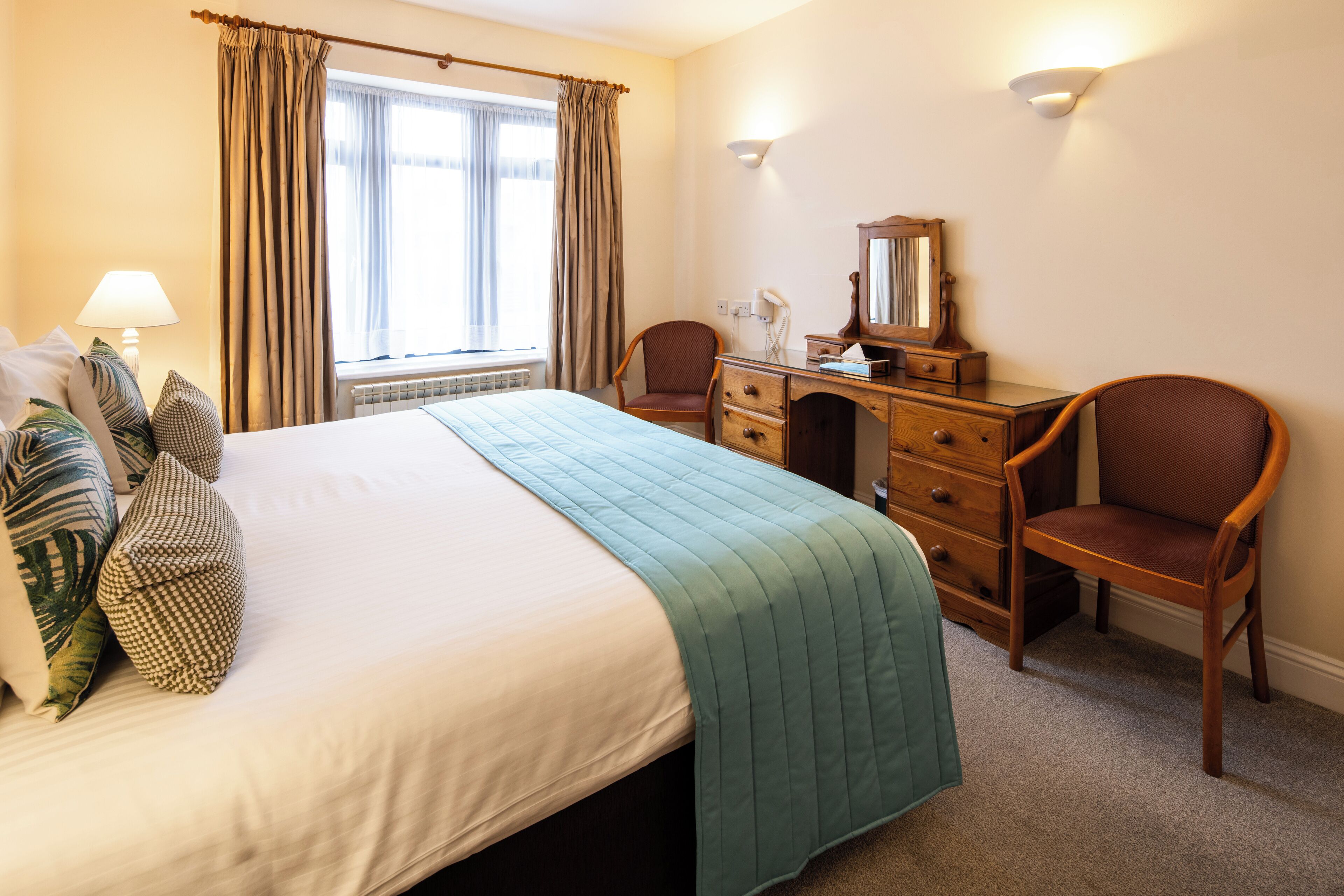 A hotel room with a bed, a desk, a chair, a lamp, and a window with curtains.
