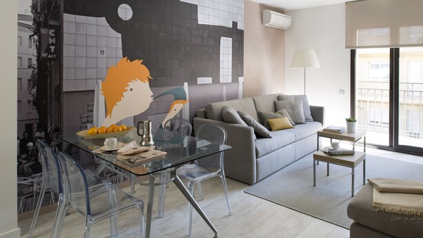 Una sala de estar moderna con una mesa de vidrio, sillas transparentes, un sofá gris con cojines y un gran mural abstracto en la pared.