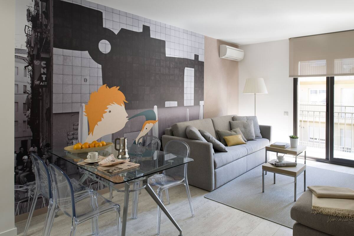 Una sala de estar moderna con una mesa de vidrio, sillas transparentes, un sofá gris con cojines y un gran mural abstracto en la pared.
