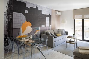 Un salon moderne avec une table à manger en verre, des chaises transparentes, un canapé gris agrémenté de coussins et une grande fresque abstraite sur le mur.