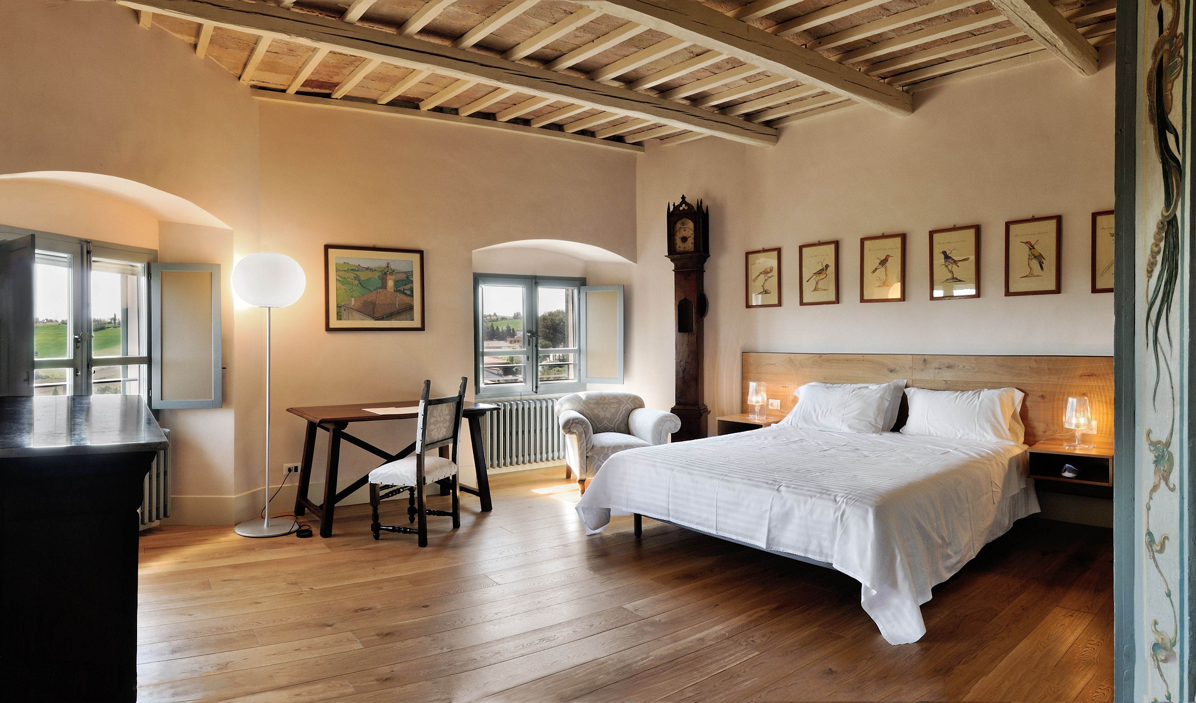 A bedroom with a wooden ceiling, a bed, a chair, a table, and a window with a view of greenery.