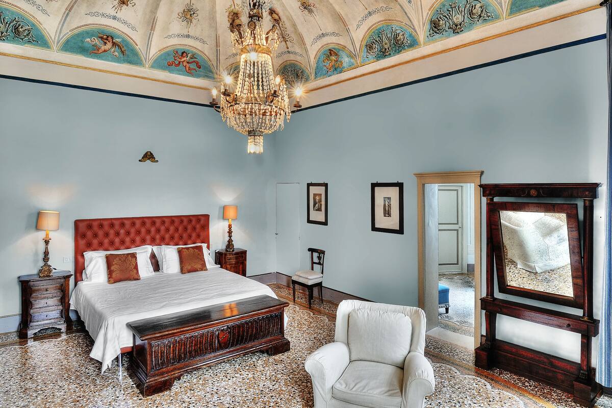 A spacious bedroom with a large bed, a chandelier, a mirror, and a chair.