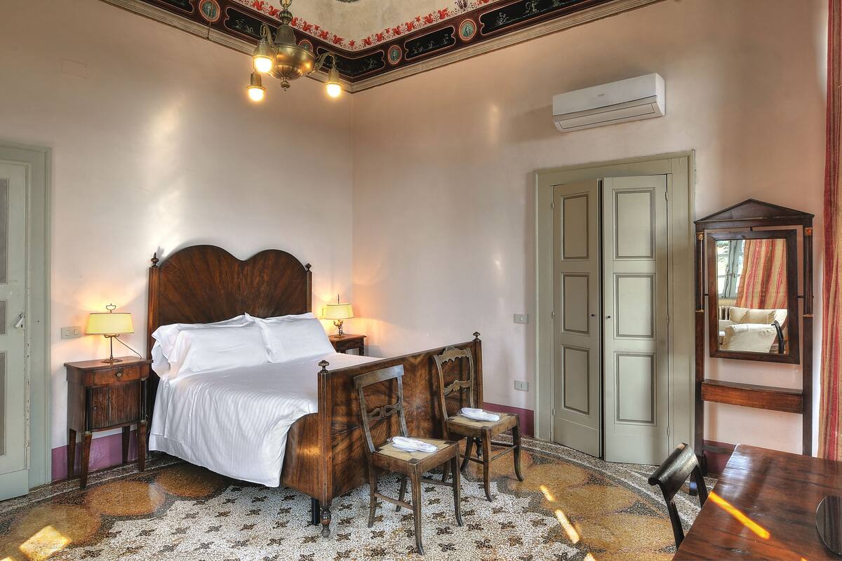 A bedroom with a wooden four-poster bed, two bedside tables with lamps, a chair, a mirror, and a door.