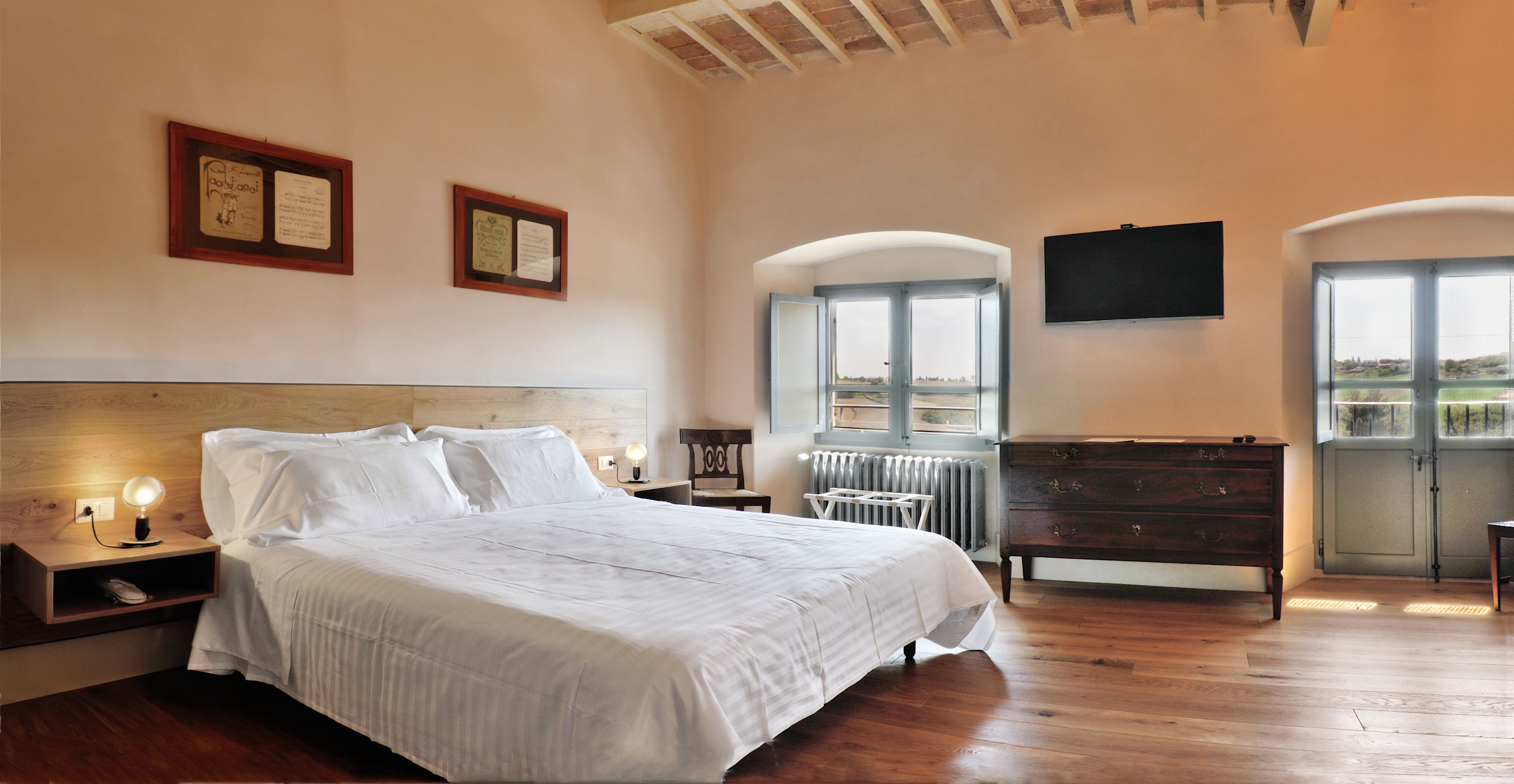 Un dormitorio con una cama grande, cabecera de madera, mesitas de noche con lámparas, un aparador, un radiador y un televisor montado en la pared.