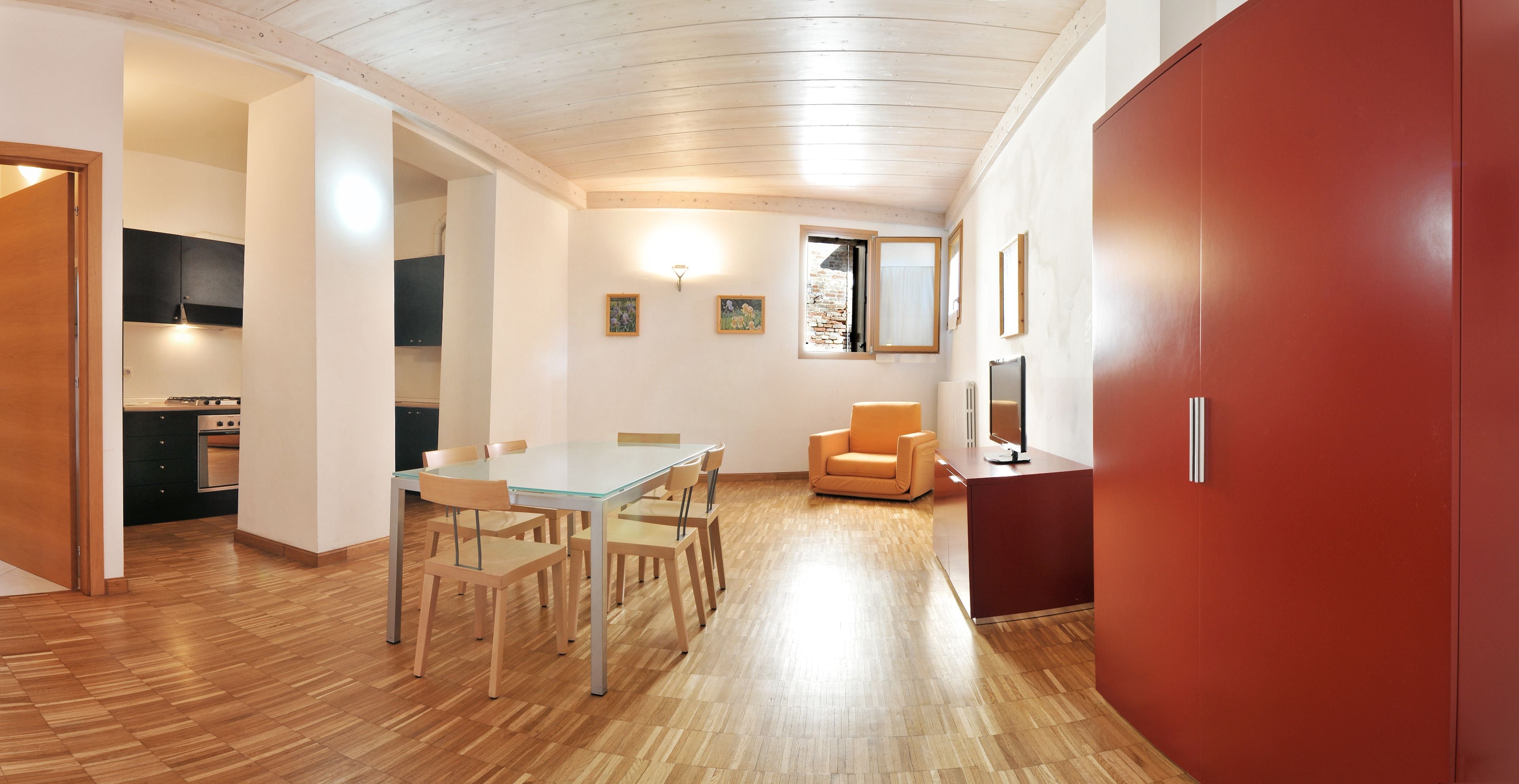 A modern kitchen with wooden flooring, a dining area with a glass table and chairs, and a red door.