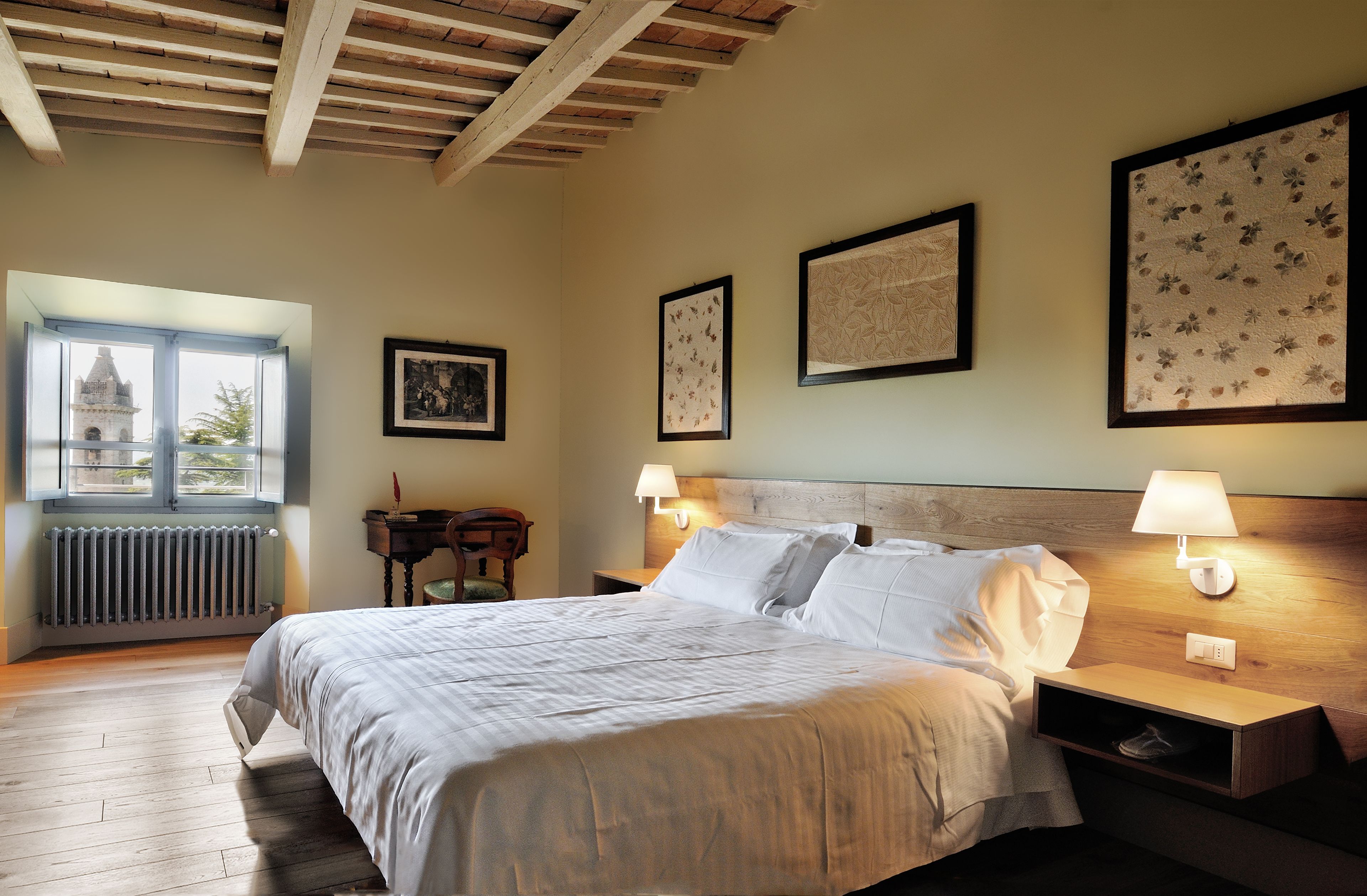 Une chambre avec un plafond en bois, un grand lit recouvert de draps blancs, deux lampes de chevet et des œuvres d’art encadrées accrochées aux murs.