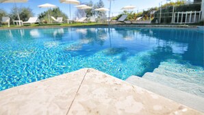 Ein Swimmingpool mit klarem, blauem Wasser, Stufen zum hineinsteigen und weiße Sonnenschirme sowie Liegestühle im Hintergrund.