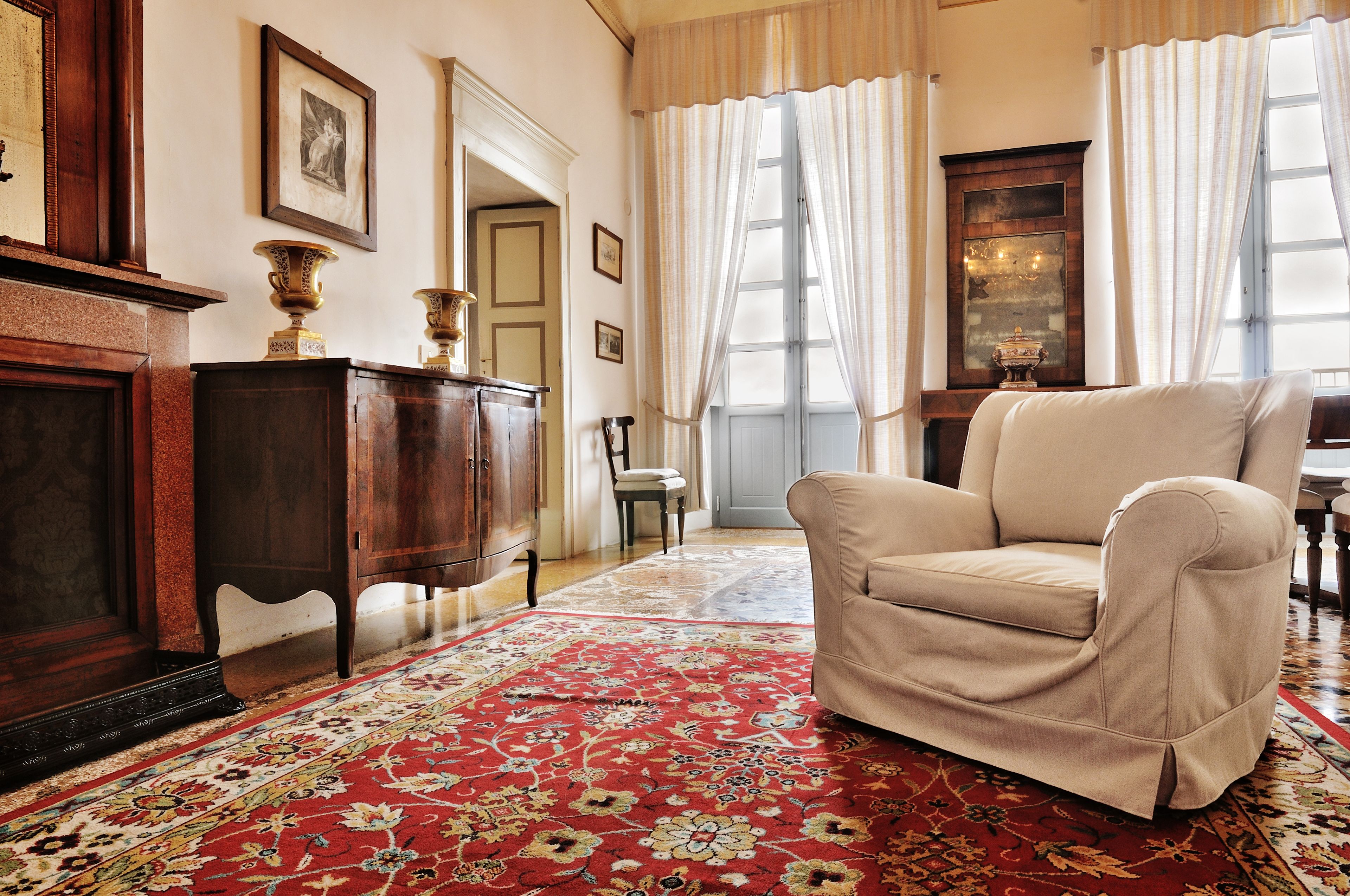 A room with a red floral carpet, a beige armchair, wooden furniture, and a large window with curtains.