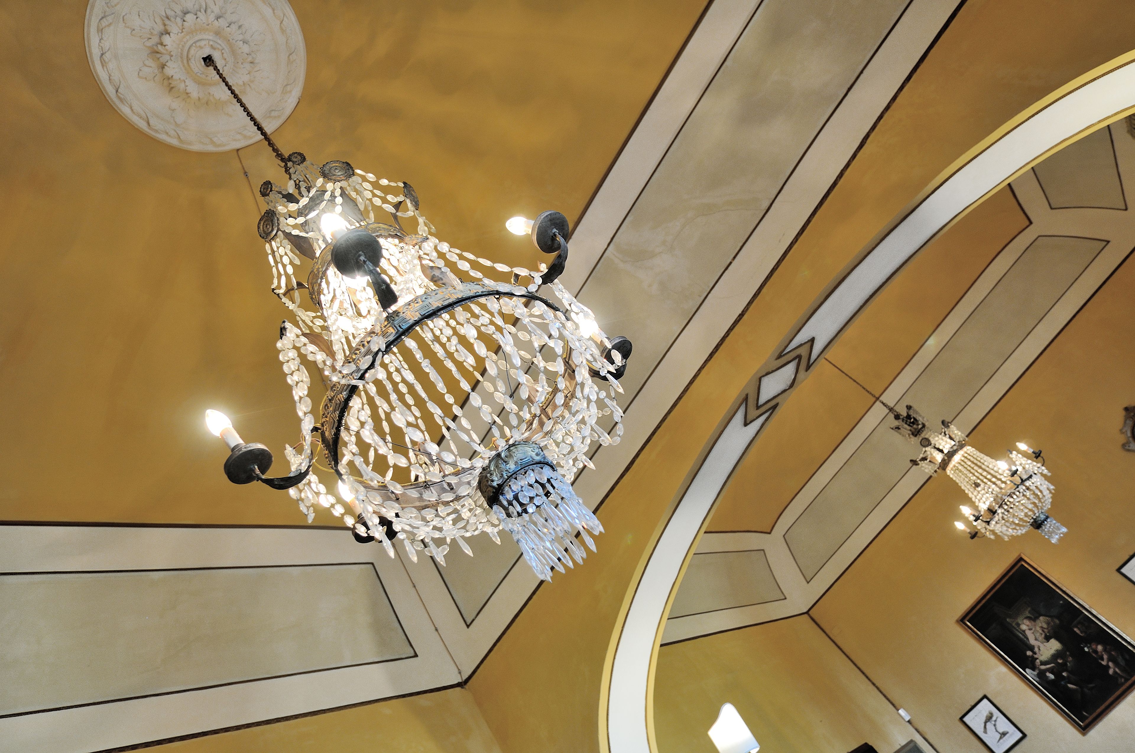 Ein prächtiger Kronleuchter mit mehreren Armen und hängenden Kristallornamenten in einem geräumigen Raum mit hoher Decke und einer Wandgestaltung im Bogenstil.