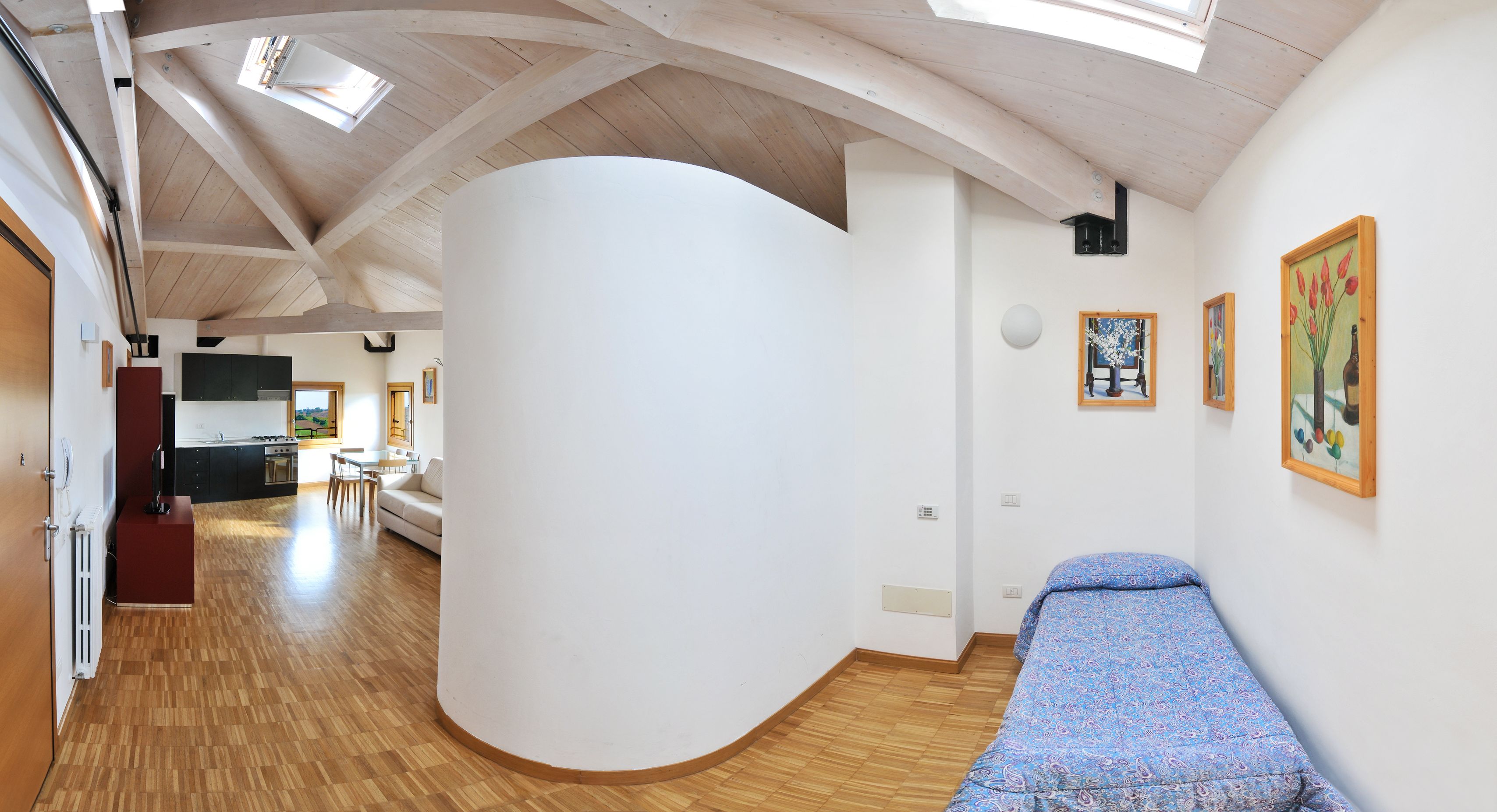 A room with a curved white wall, wooden flooring, a bed with a patterned bedspread, and a dining area with a table and chairs.