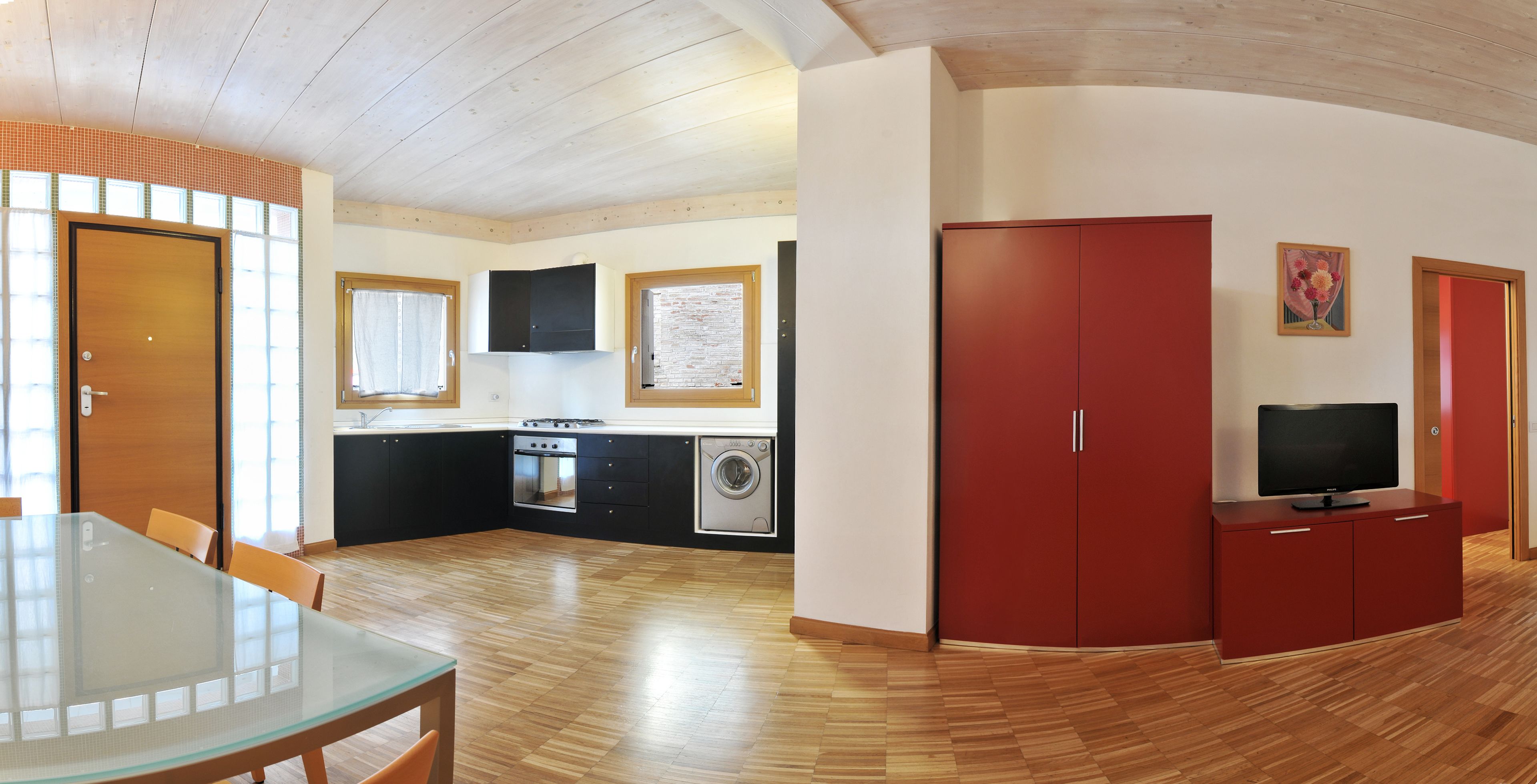 Cocina compacta con lavadora, zona de comedor con mesa de vidrio y un armario rojo.