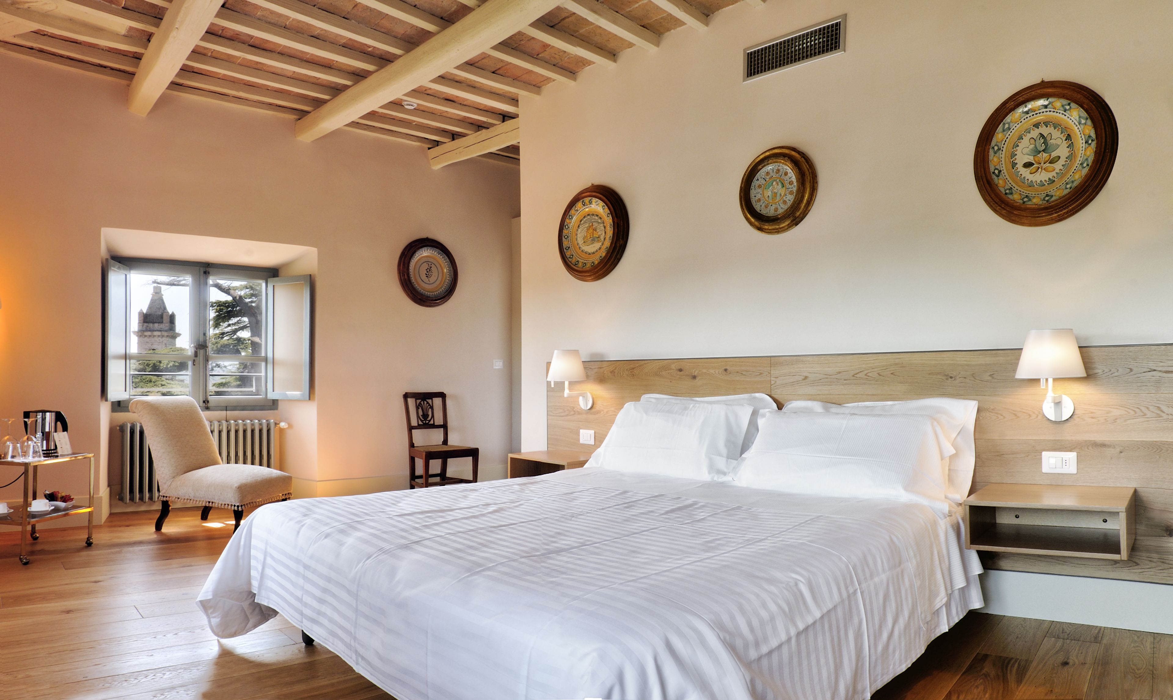 A bedroom with a large bed, wooden headboard, bedside tables with lamps, a chair, a radiator, and a window with a view.