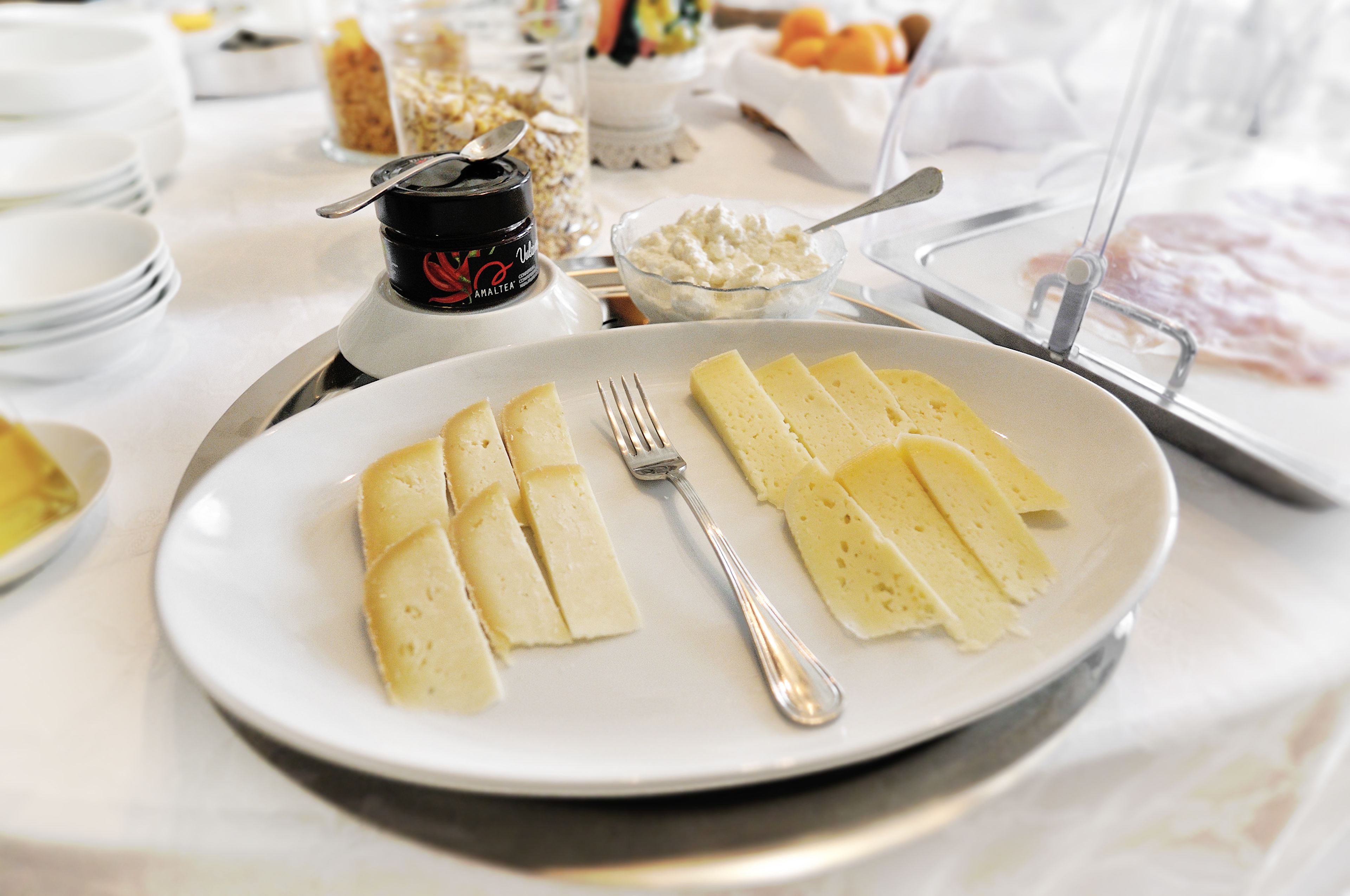 Un piatto di formaggio a fette con una forchetta su un piatto bianco, circondato da diversi cibi e condimenti su un tavolo.