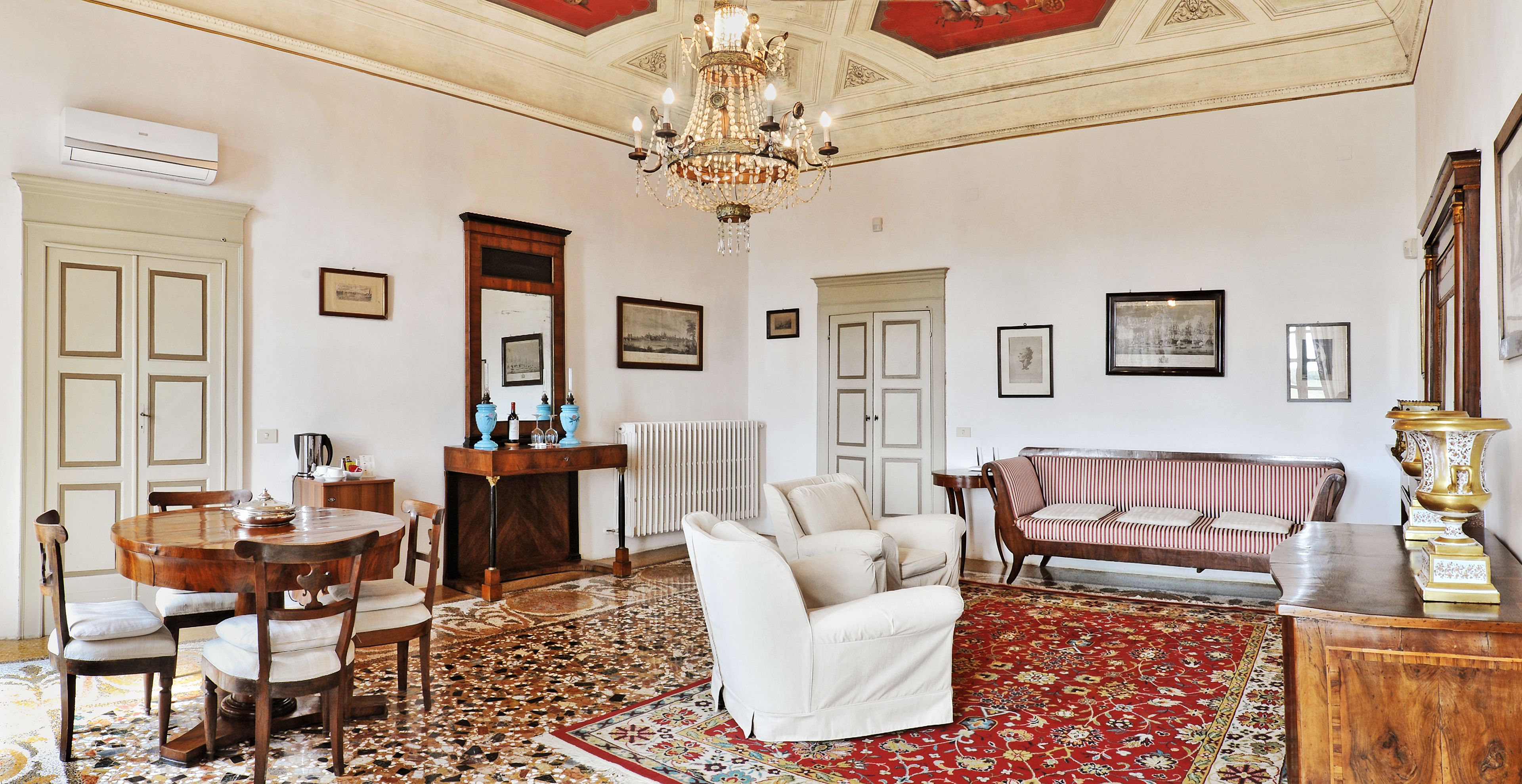 Un'ampia stanza con un tavolo da pranzo rotondo in legno, sedie imbottite bianche, un divano a righe rosse e bianche e un grande lampadario.