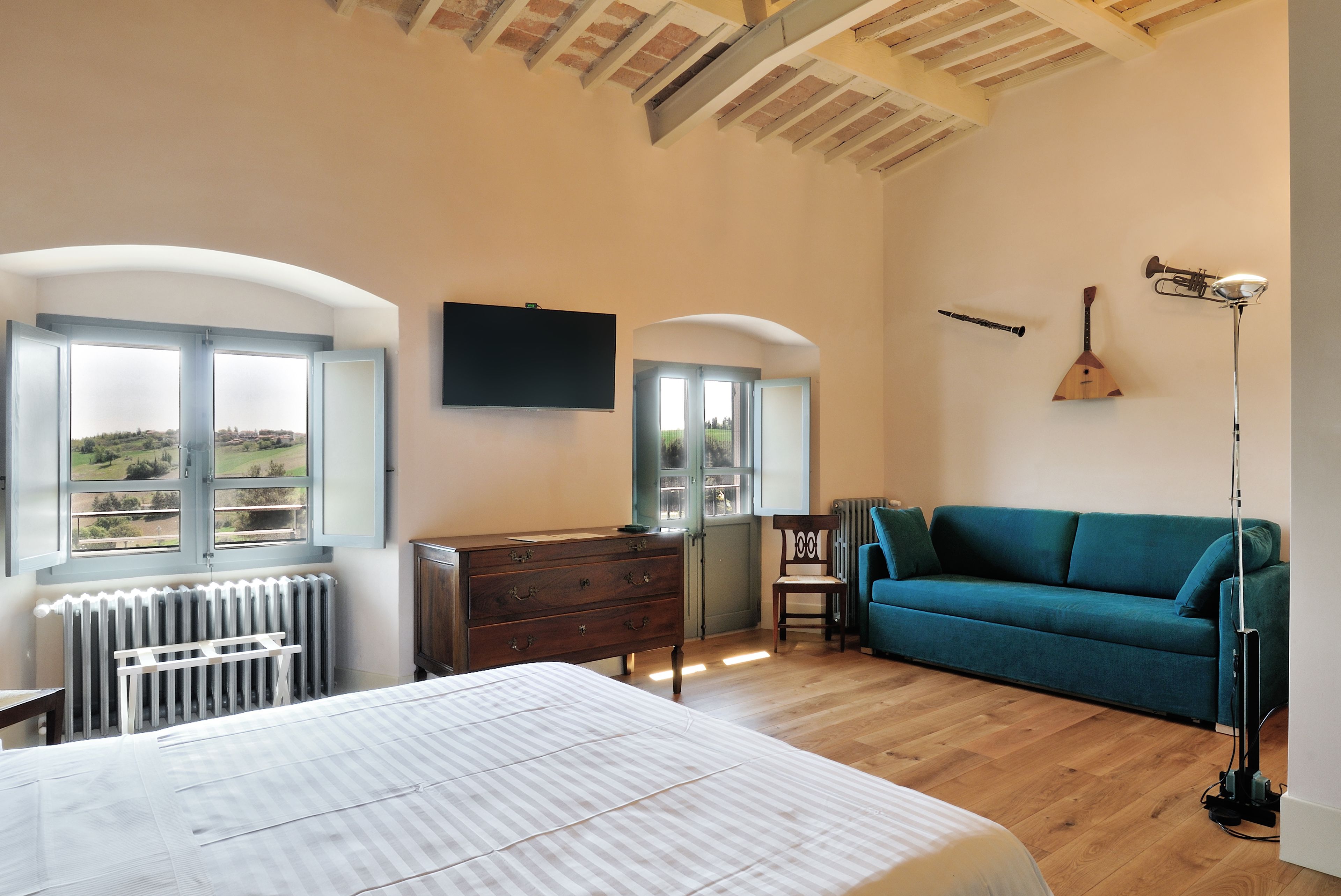 A bedroom with a bed, a dresser, a blue sofa, a chair, a radiator, a TV, and a view of the outdoors.