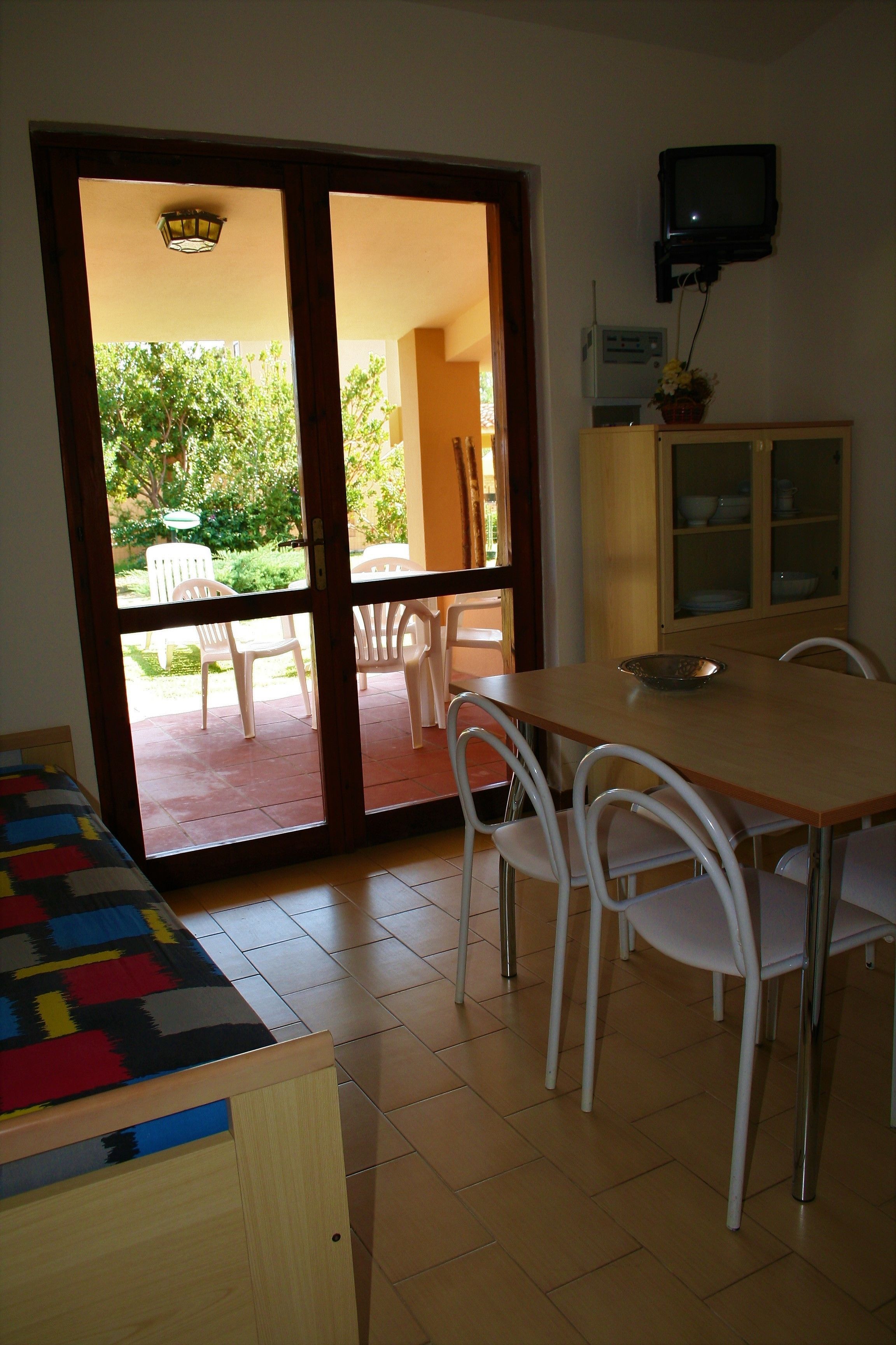 A room with a wooden table, white chairs, a television mounted on the wall, and a sliding glass door leading to an outdoor area with chairs and greenery.