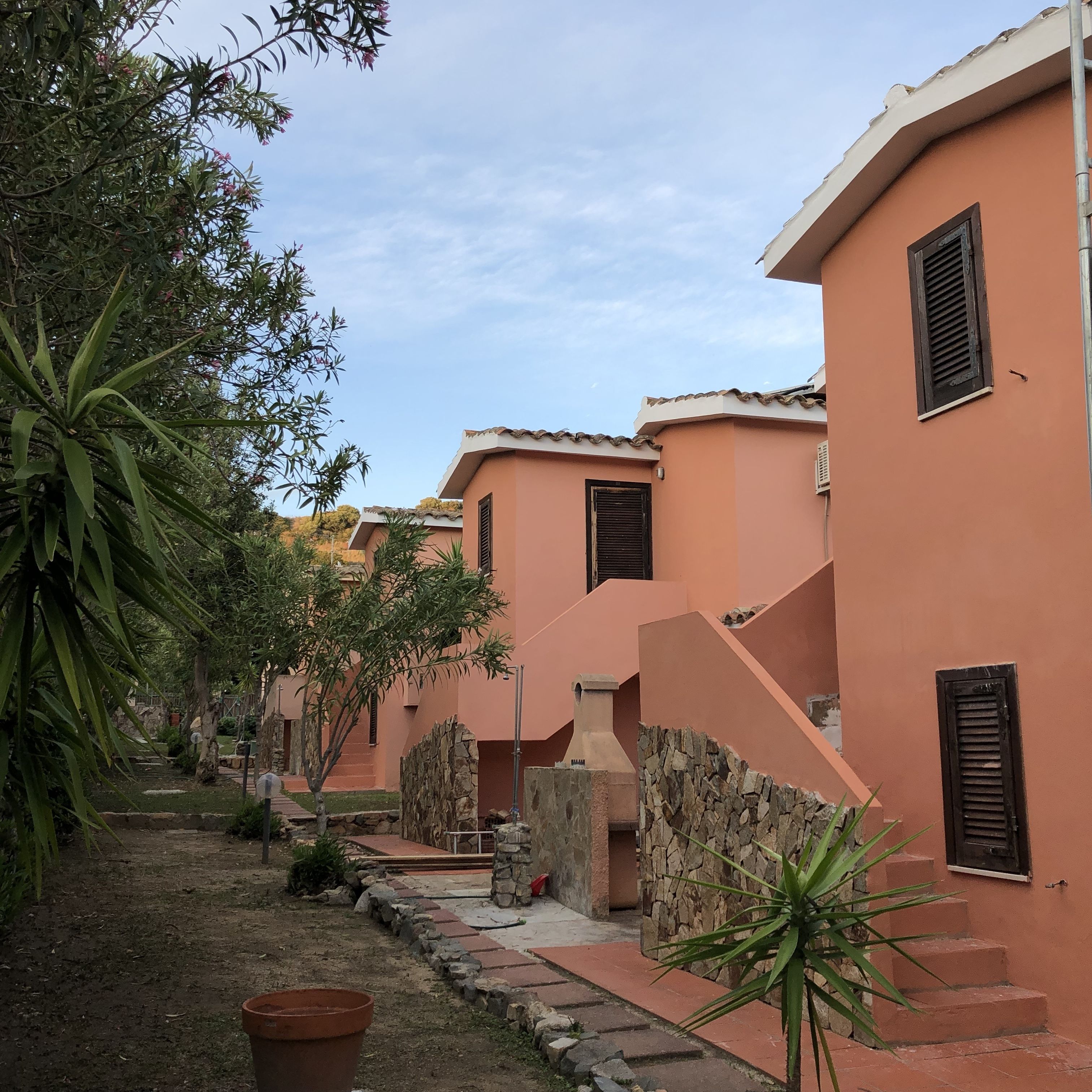 A row of pink buildings with stone walls and wooden shutters, a paved pathway, and potted plants.