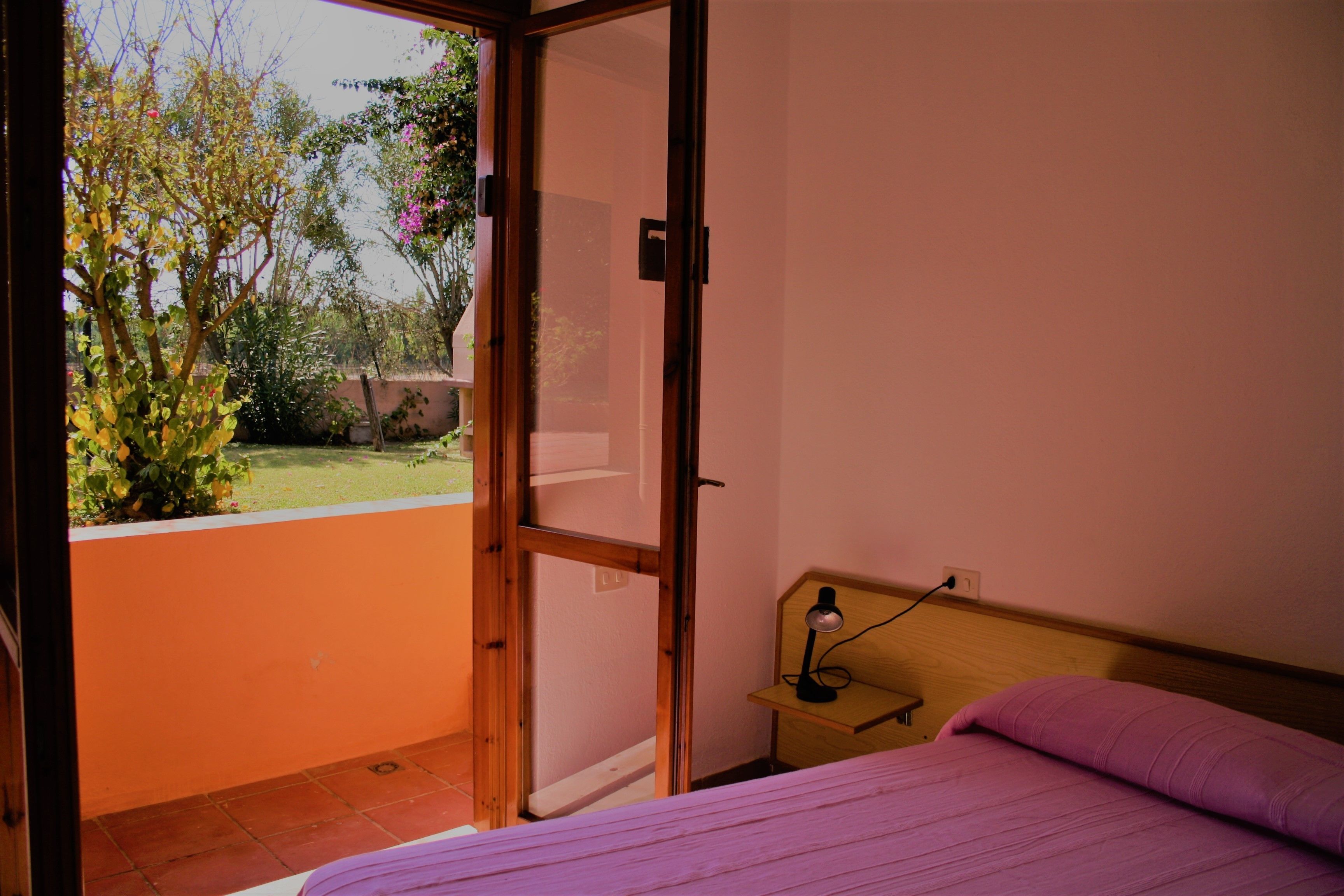 Une chambre à coucher avec un lit, une table de chevet et une vue sur un jardin, visible à travers une porte ouverte.