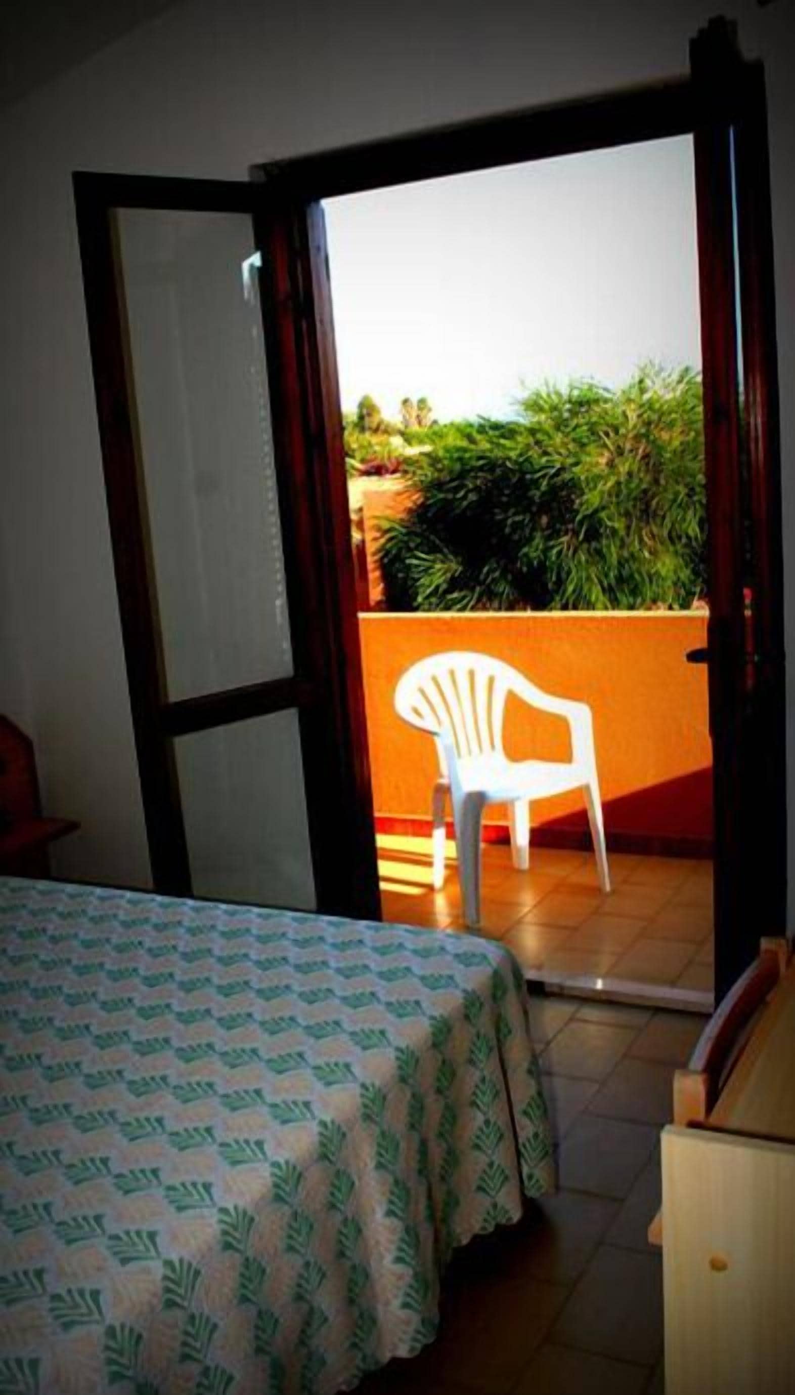 Una habitación con una cama, una silla blanca y una puerta abierta que da a un balcón con plantas.