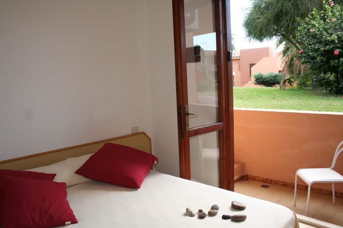Quarto com cama, almofadas vermelhas, cadeira branca e varanda com vista para um jardim e um edifício.