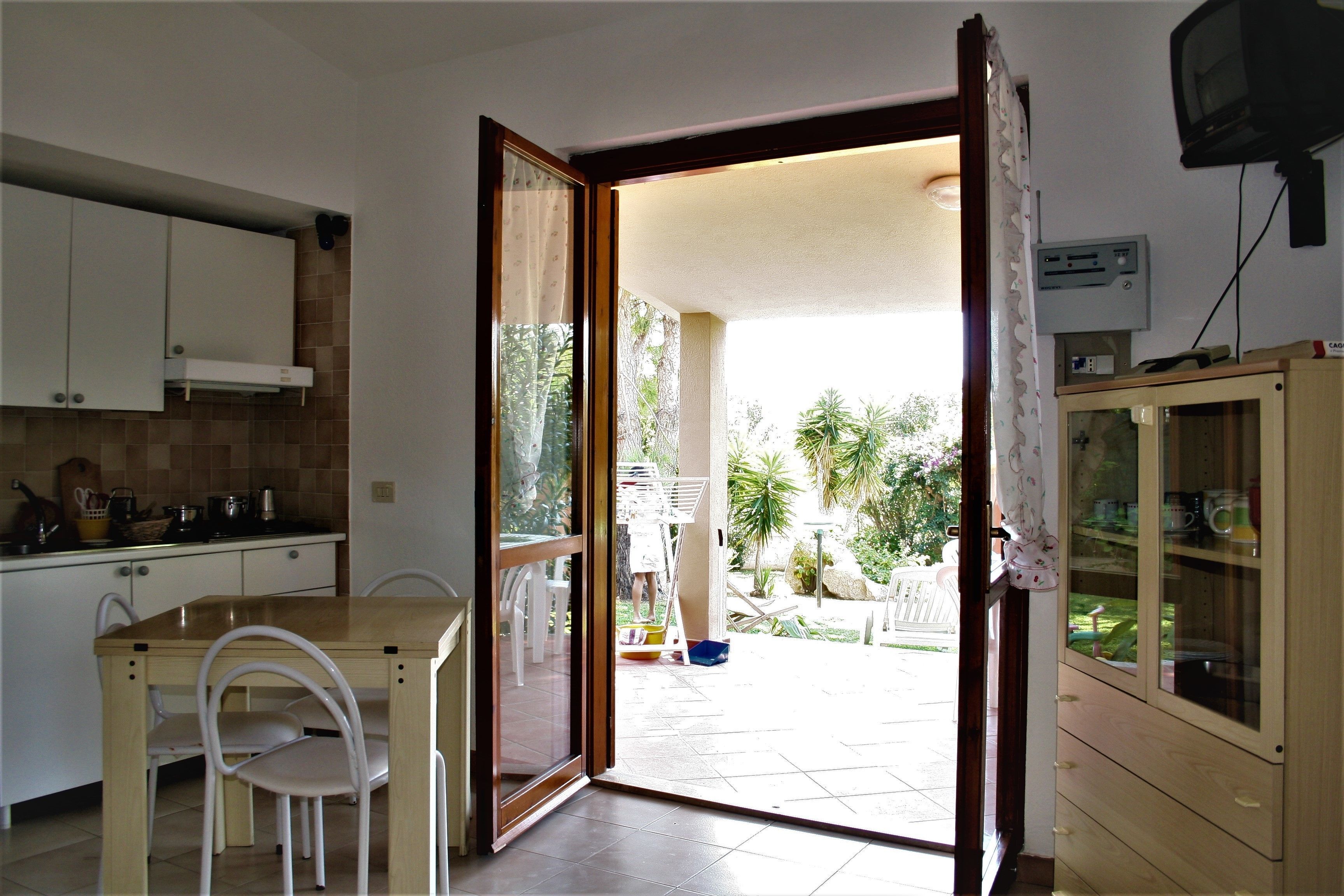 A kitchen with white cabinets, a wooden table, and chairs. There is a sliding glass door leading to an outdoor area with a garden.