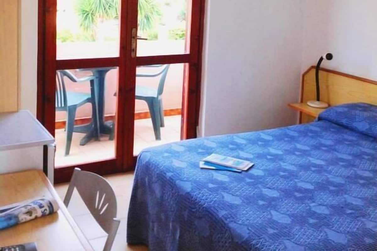 Quarto com cama, escrivaninha com cadeira e porta de vidro deslizante para área externa com mesa e cadeiras.