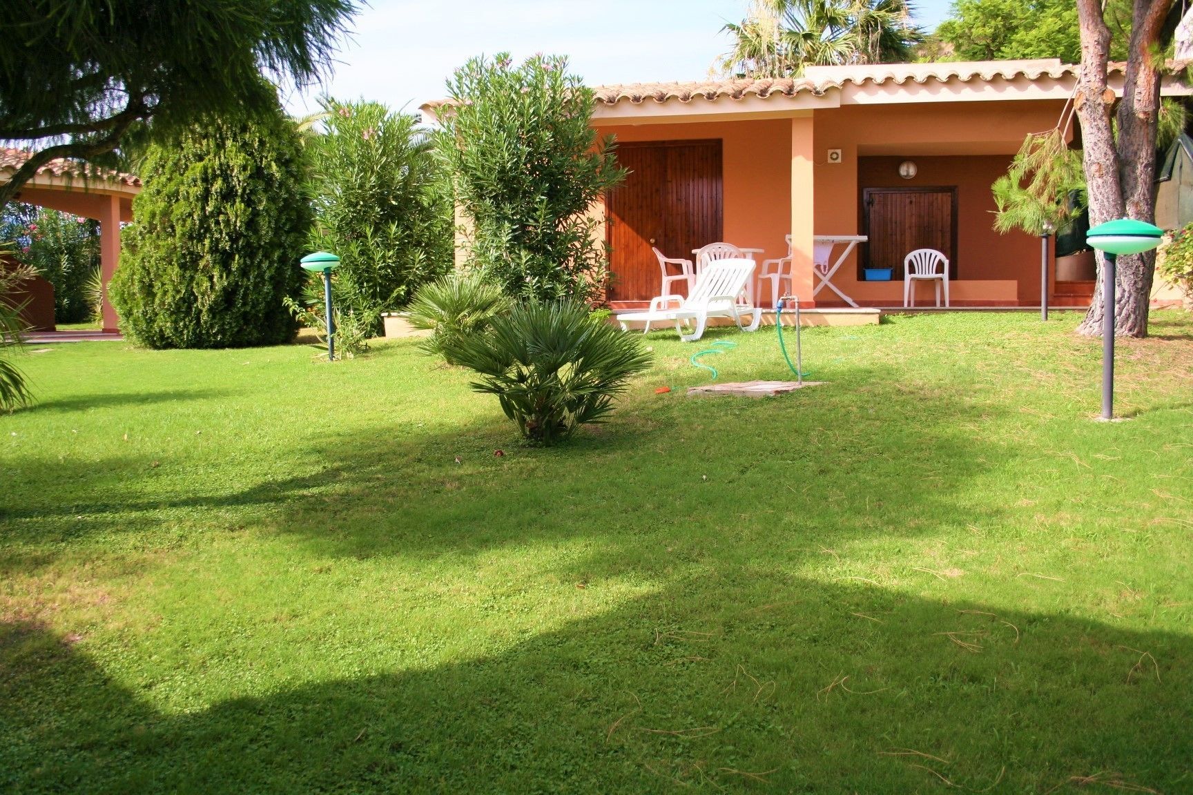 A well-maintained garden with a lawn, a small palm tree, and a patio area with white chairs and a table.