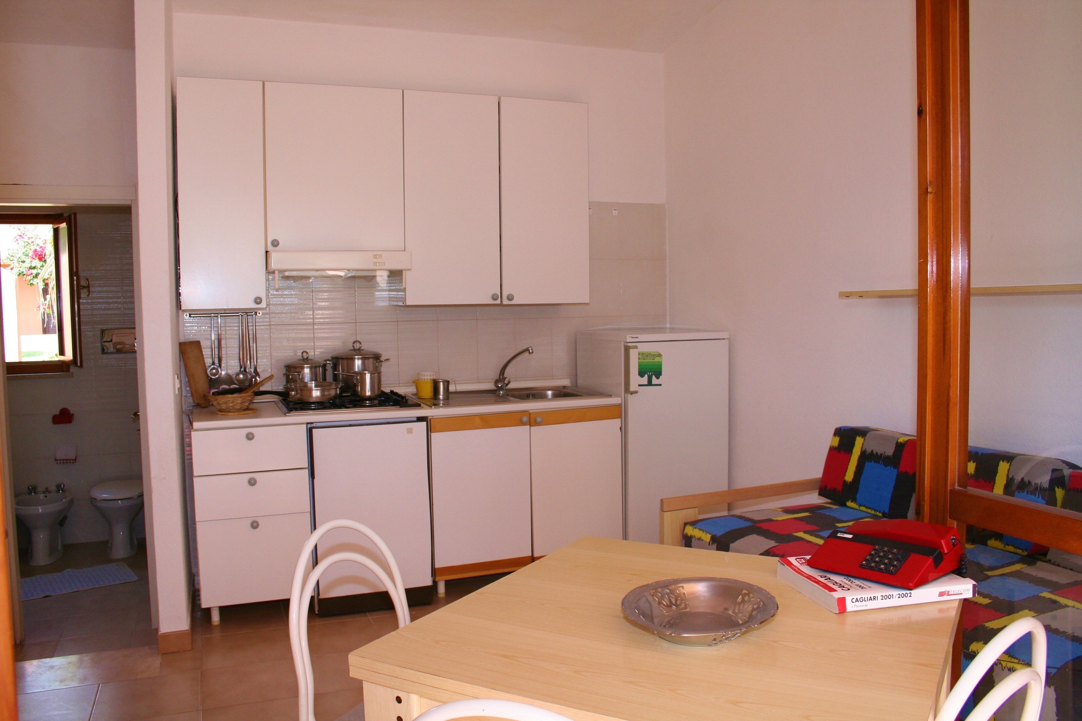 Una cocina compacta con armarios blancos, un lavamanos y un refrigerador pequeño. Hay una mesa de comedor con un tazón de metal y un libro. Un sofá colorido se ve al fondo.