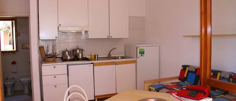 Una cocina compacta con armarios blancos, un lavamanos y un refrigerador pequeño. Hay una mesa de comedor con un tazón de metal y un libro. Un sofá colorido se ve al fondo.