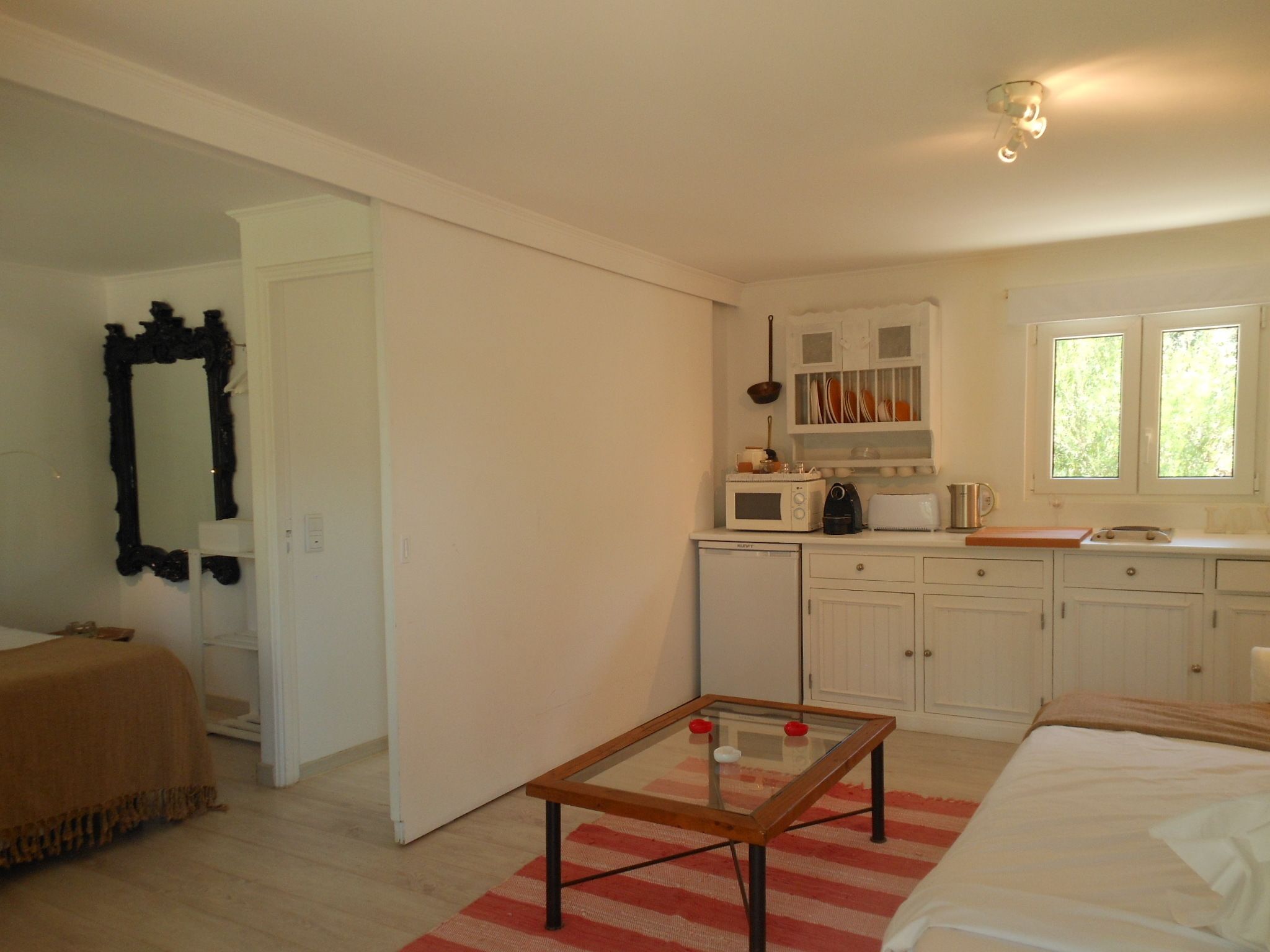 A compact, well-lit room with a kitchenette, a bed, a glass-top table, and a mirror.