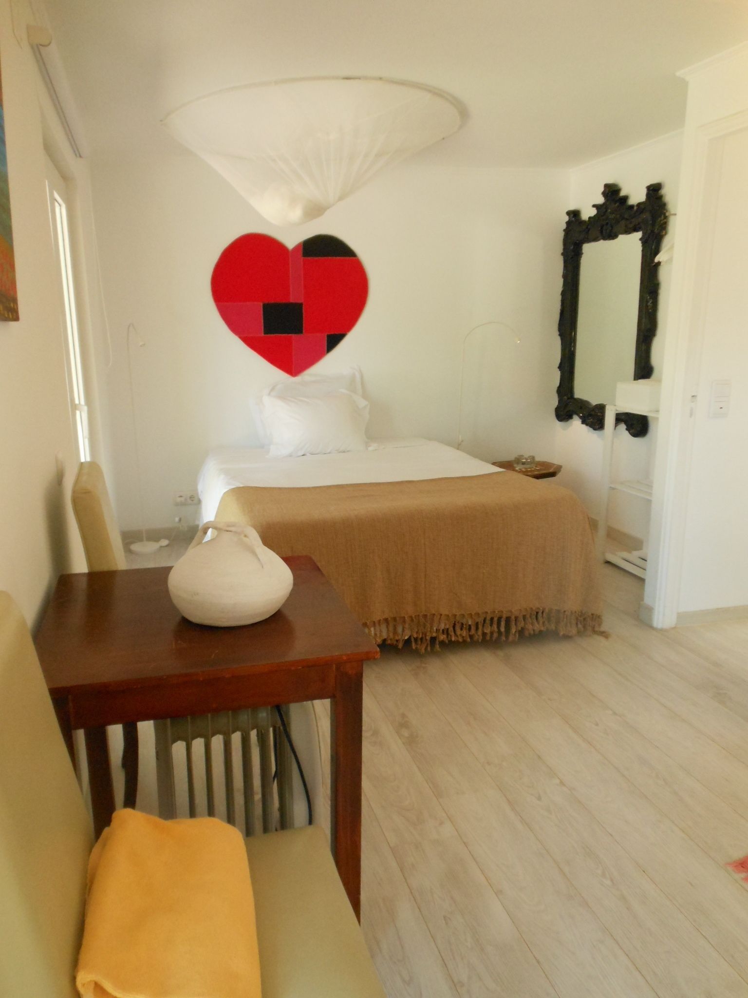 A hotel room with a bed, a table with a vase, a heart-shaped decoration on the wall, and a mirror.