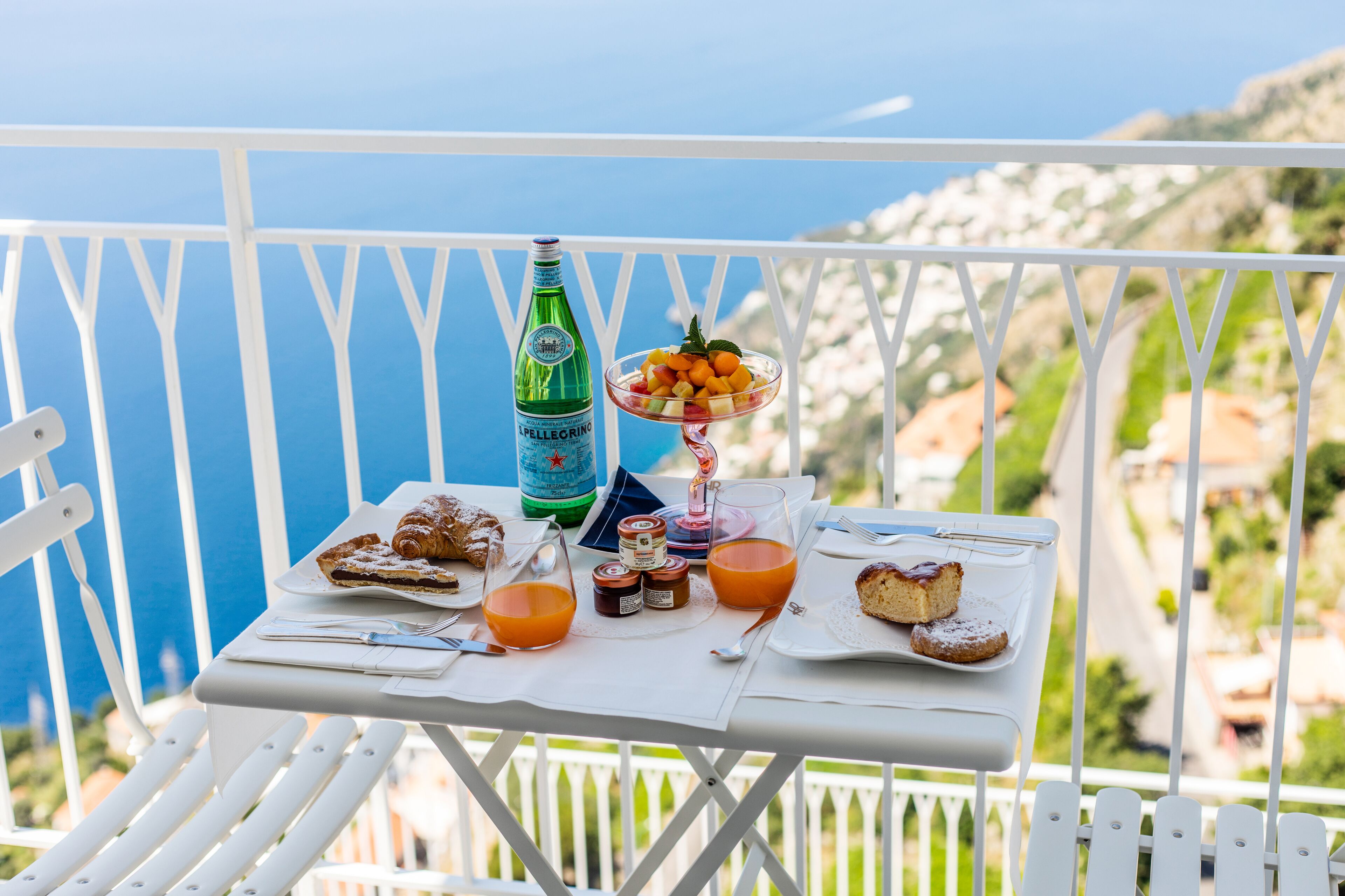 Une table avec des aliments pour le petit-déjeuner, une bouteille et un verre de jus d’orange, sur un balcon donnant sur la mer.