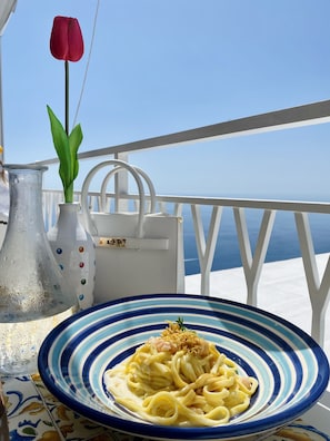 A table with a plate of pasta, a vase with a red flower, and a white handbag on a balcony with a view of the sea.
