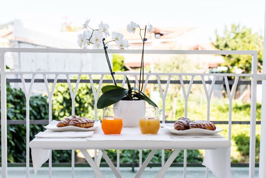 A white table with two plates of pastries and two glasses of orange juice on a balcony with a white railing and greenery in the background.