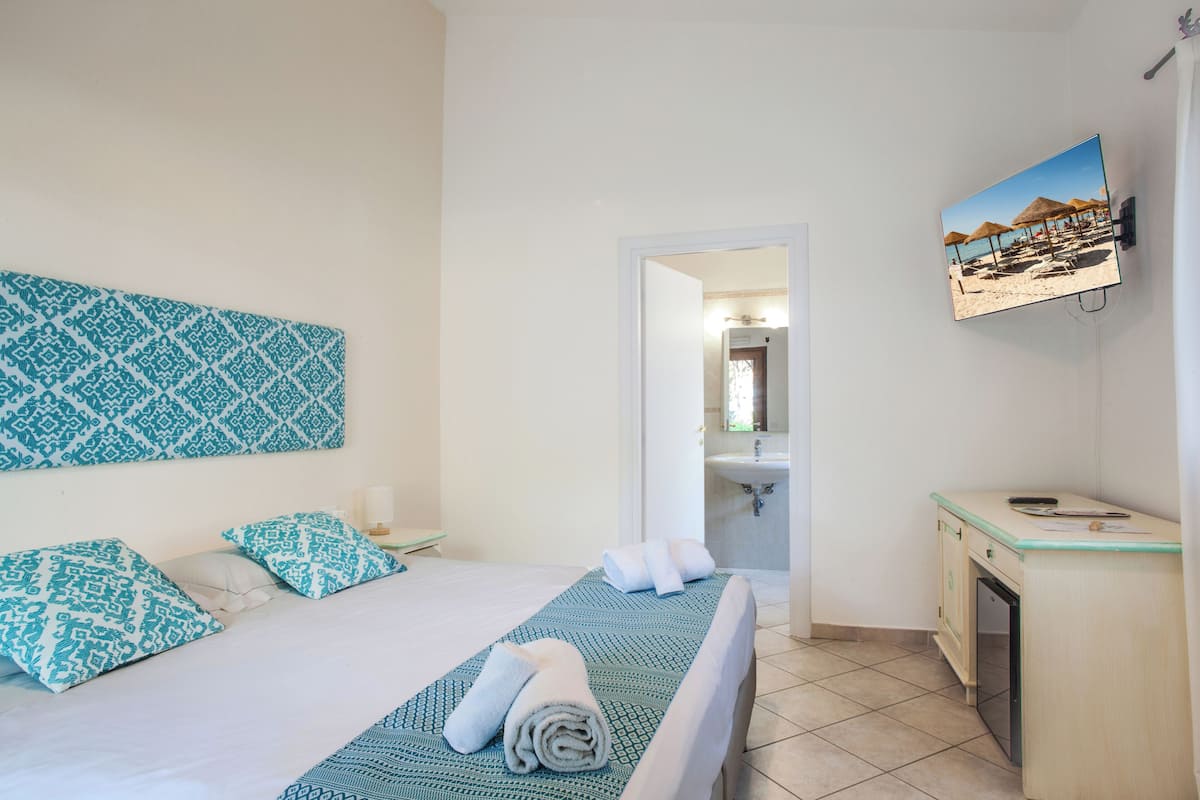 Ein Hotelzimmer mit Bett, Schreibtisch, Badezimmer und Fernseher, der eine Strandansicht zeigt.