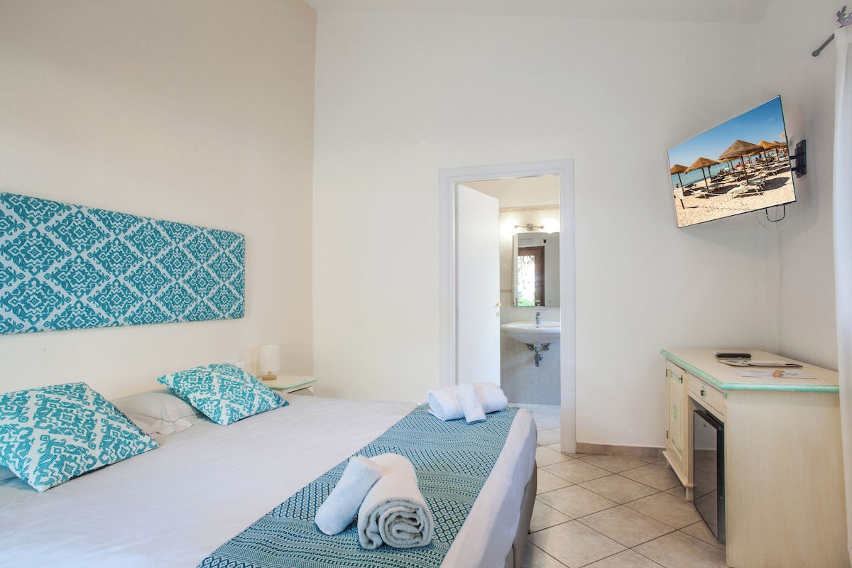 A hotel room with a bed, a desk, a bathroom, and a wall-mounted TV displaying a beach scene.