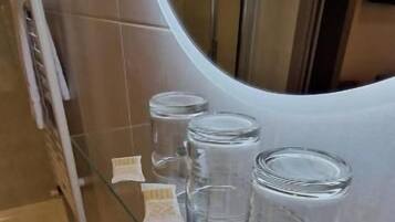 Um banheiro com prateleira de vidro, copos e pequenas garrafas, pia branca com torneira cromada e um espelho grande refletindo o ambiente.