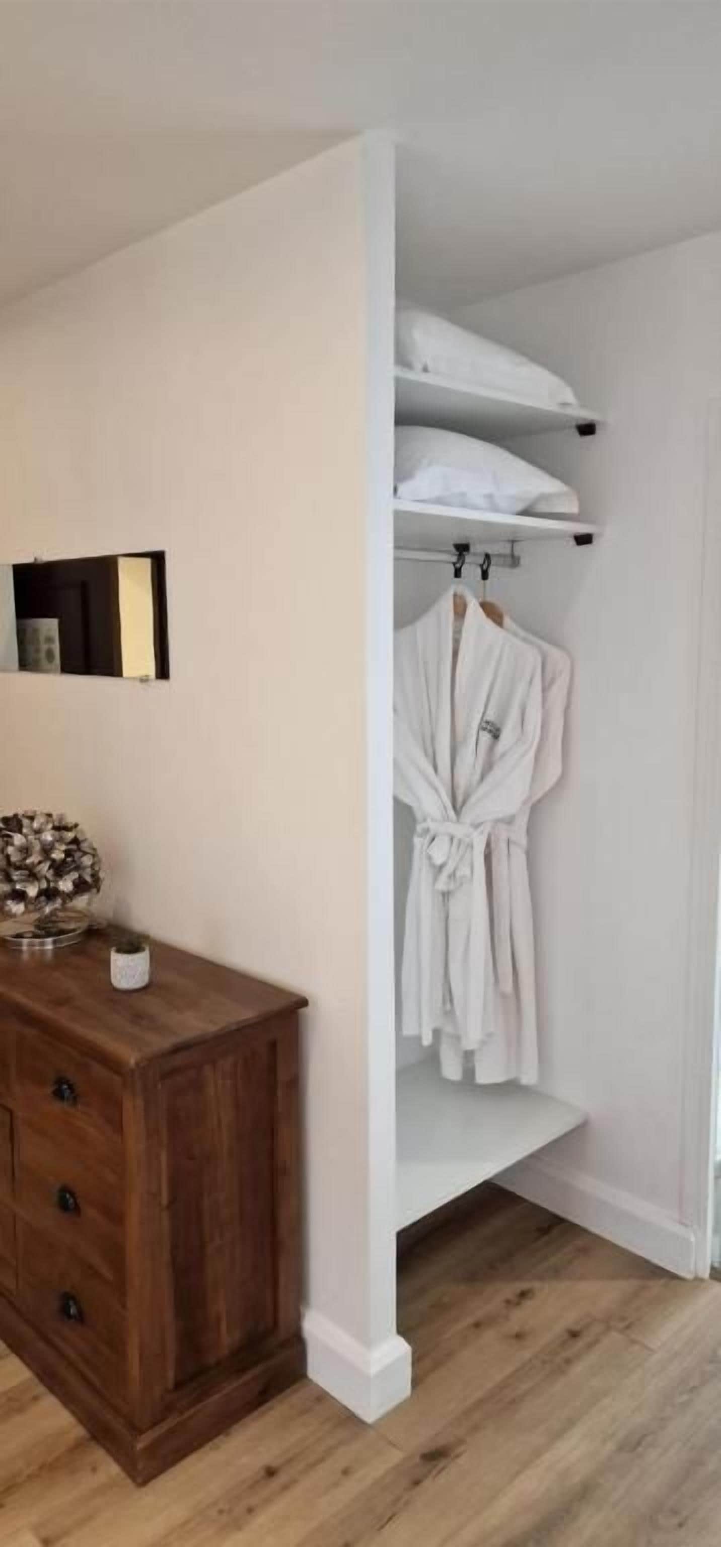 A room with a wooden dresser, a white robe hanging on a rack, and a shelf with pillows.