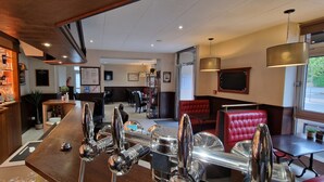 A bar area with a polished bar counter and chrome fixtures, a seating area with red leather chairs, and a view through a large window. - Hôtel Le Bellevue (Guer)