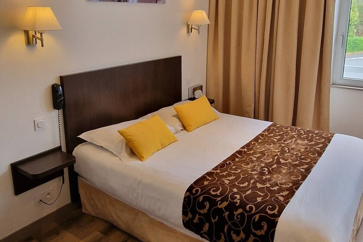 Una habitación de hotel con una cama, lámparas de noche, una mesita de noche, una silla y una ventana con cortinas.