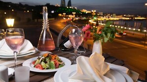 Taula ben posada per a sopar amb vasos de vi, una ampolla i un plat de amanida, amb vista a un paisatge urbà a la posta de sol.
