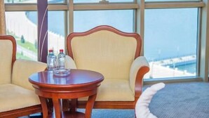 Habitació d'hotel amb un llit, dues cadires, una taula amb una làmpada i vistes a un paisatge urbà a través de grans finestres.