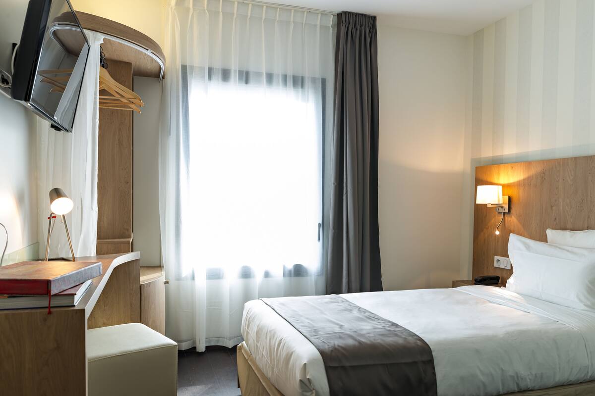 A hotel room with a bed, a desk with a lamp, a TV, and a window with curtains.