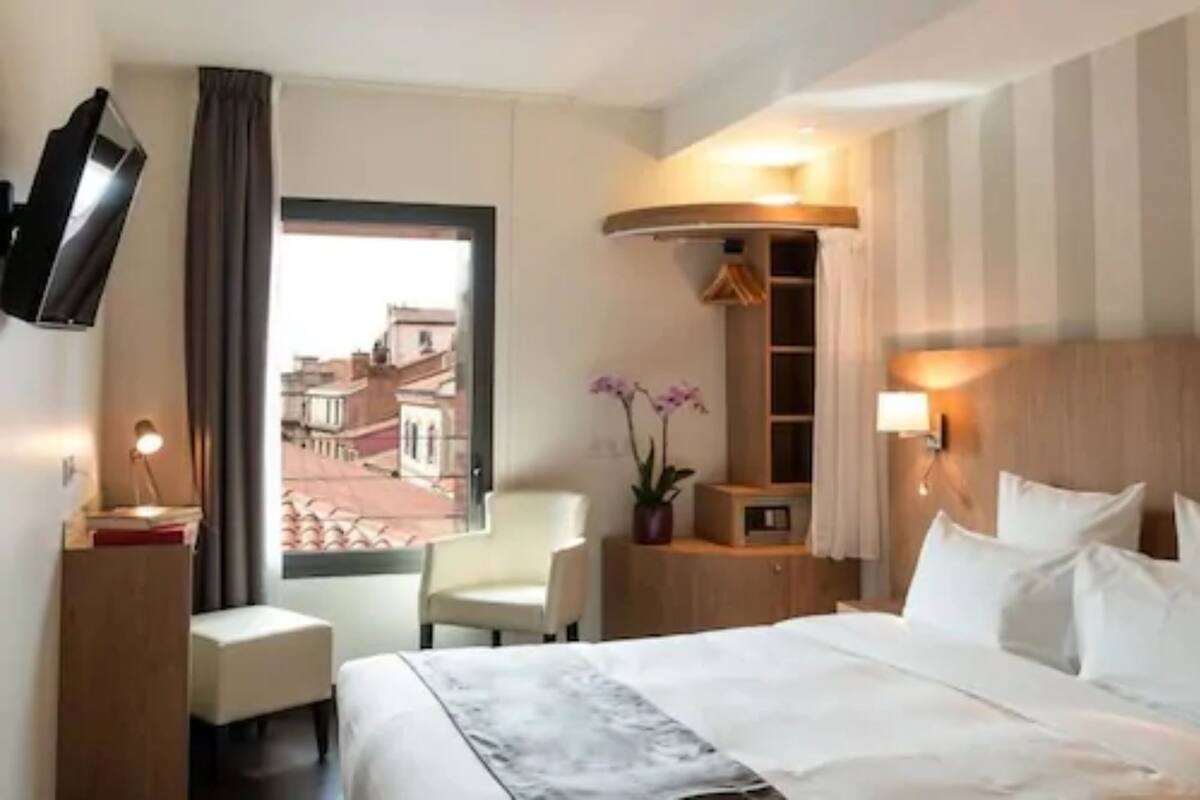 A hotel room with a bed, a desk, a chair, a TV, a window with a view of buildings, and a wall-mounted shelf with a plant.
